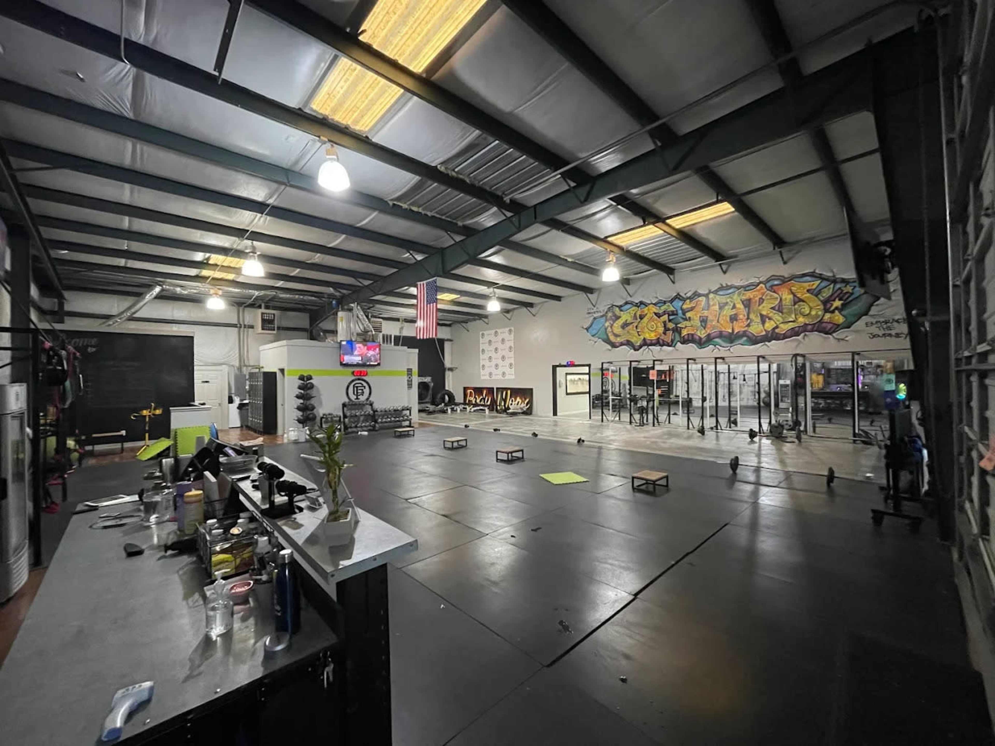 The image shows a spacious gym with exercise equipment, large open areas, and a colorful mural on the wall.