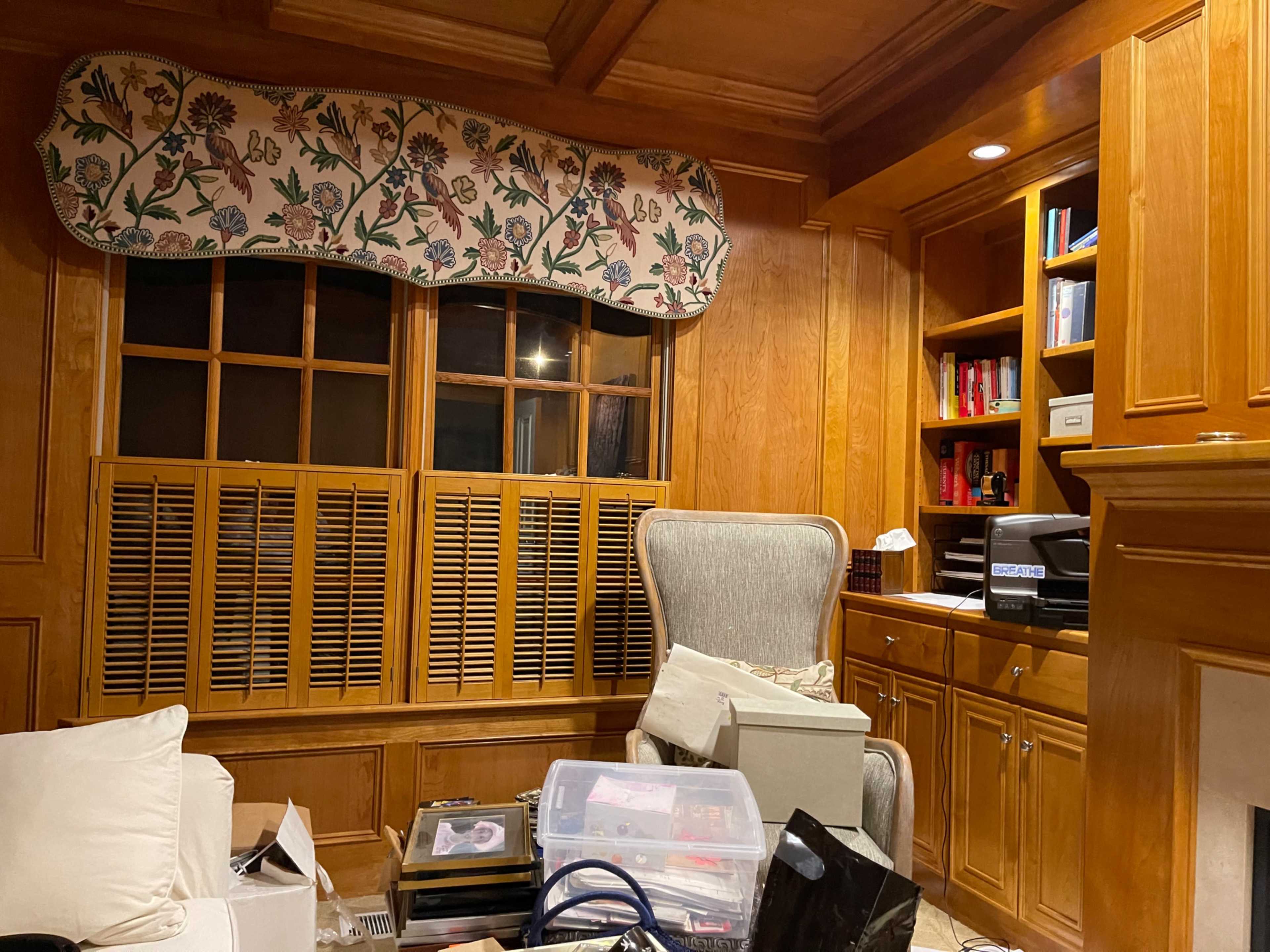 A cluttered room with wooden paneling, a large armchair, a window with a patterned valance, and shelves filled with books and boxes.