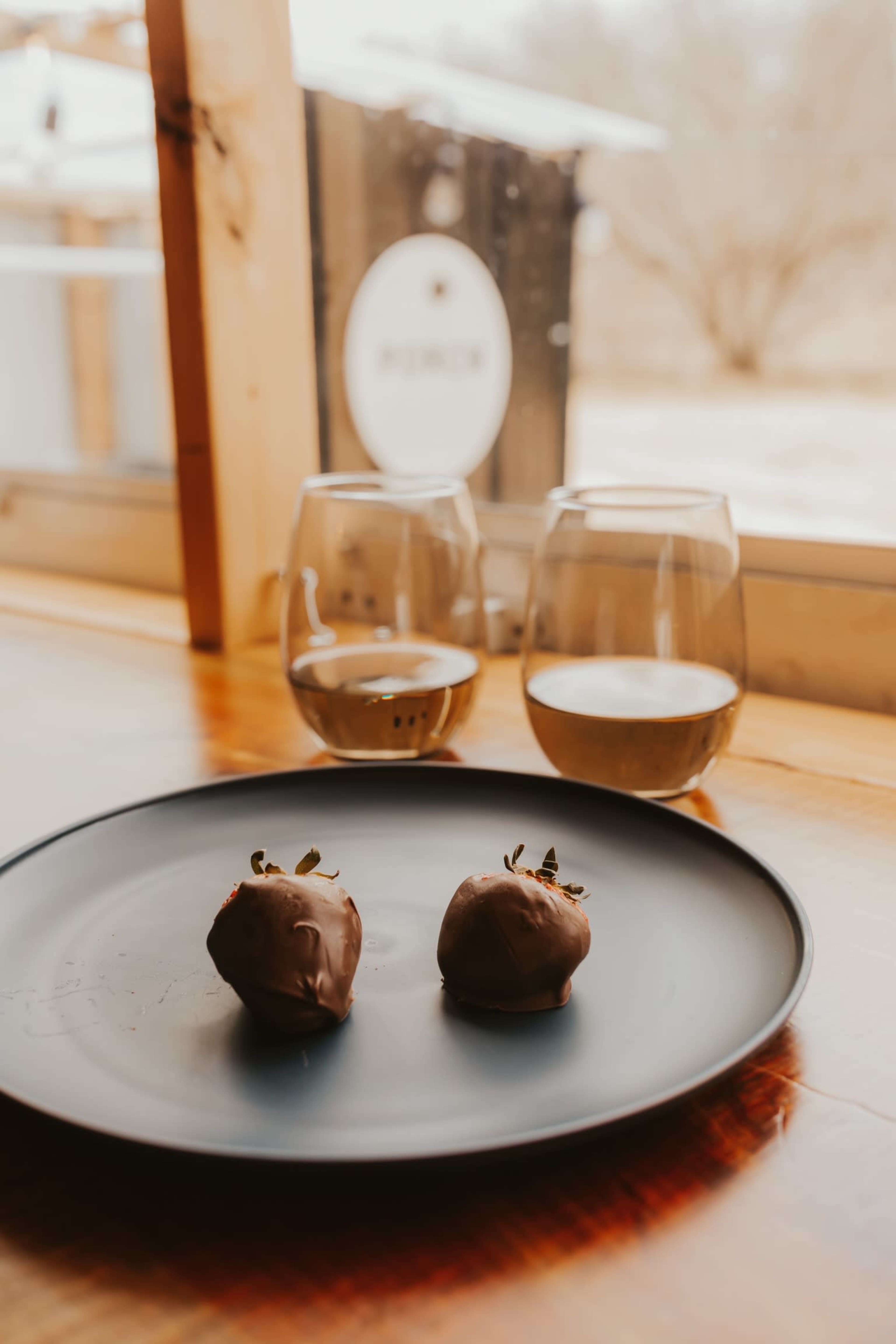 A black plate with two chocolate-covered truffles sits on a wooden table beside two glasses of white wine, with a blurred outdoor scene visible through the window.