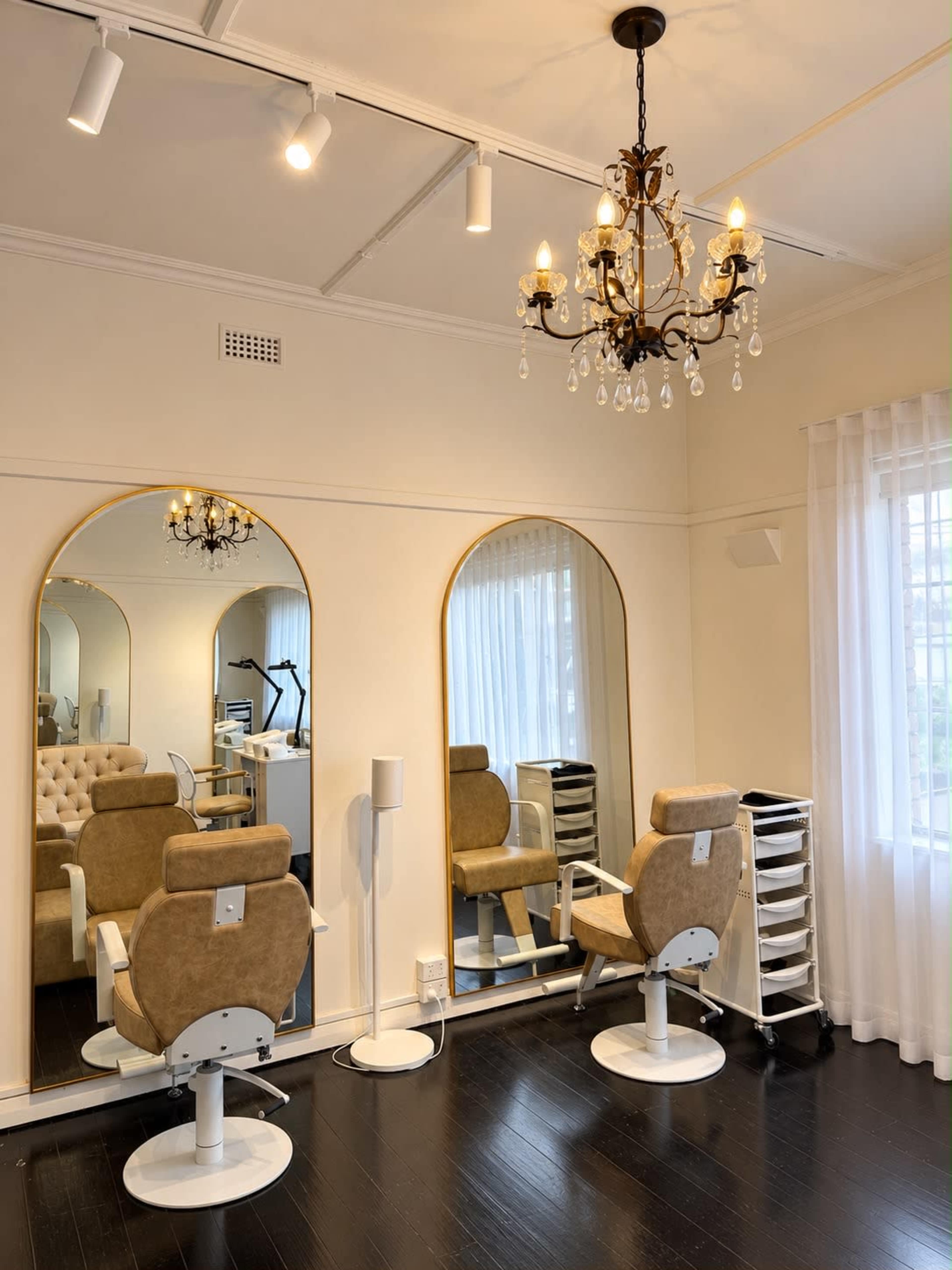 The image shows a well-lit salon interior featuring two styling chairs, large mirrors, and a chandelier, with natural light coming through a window.