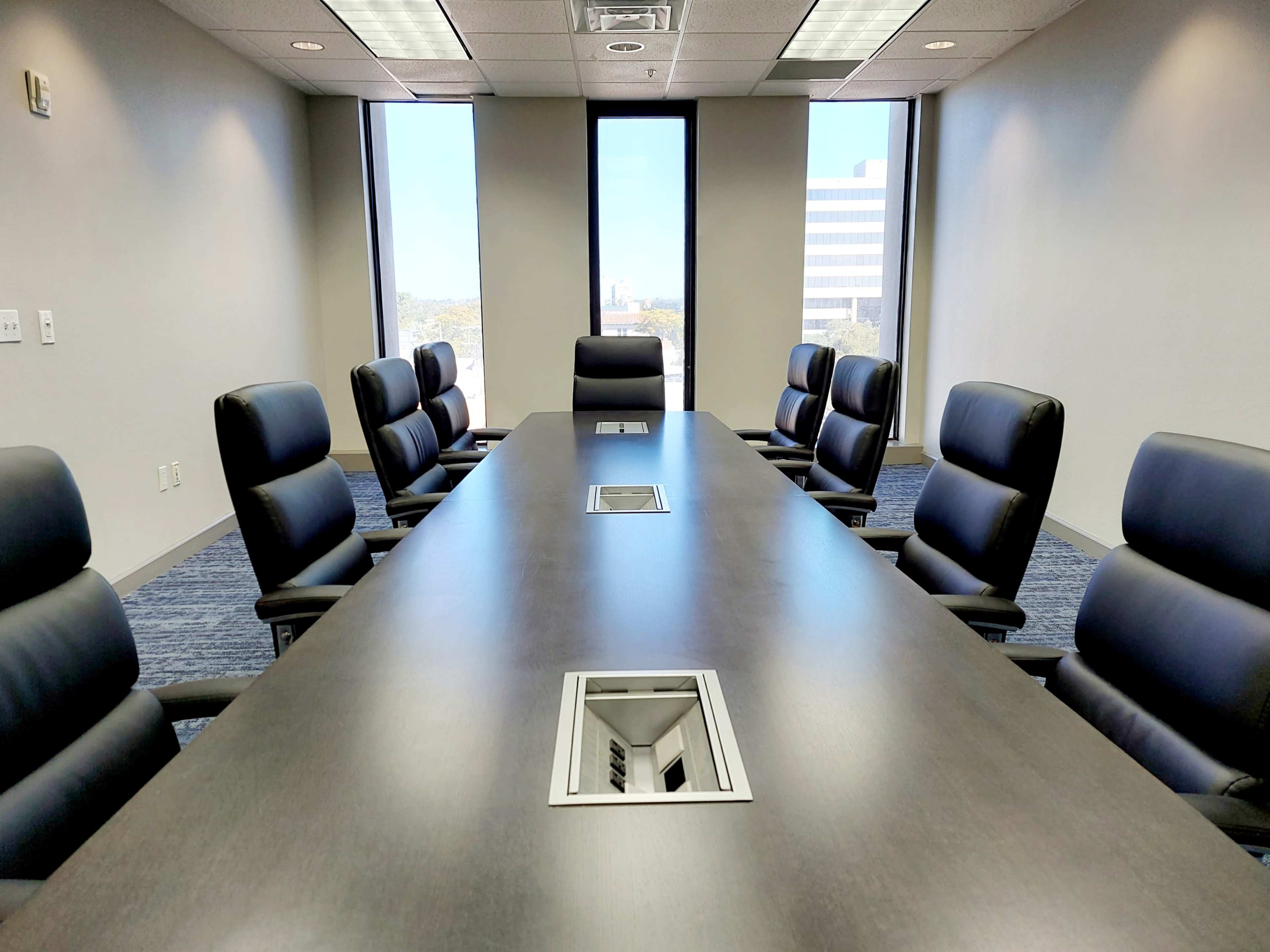 A modern conference room features a long dark table surrounded by eight leather chairs, with large windows providing a view of the cityscape.