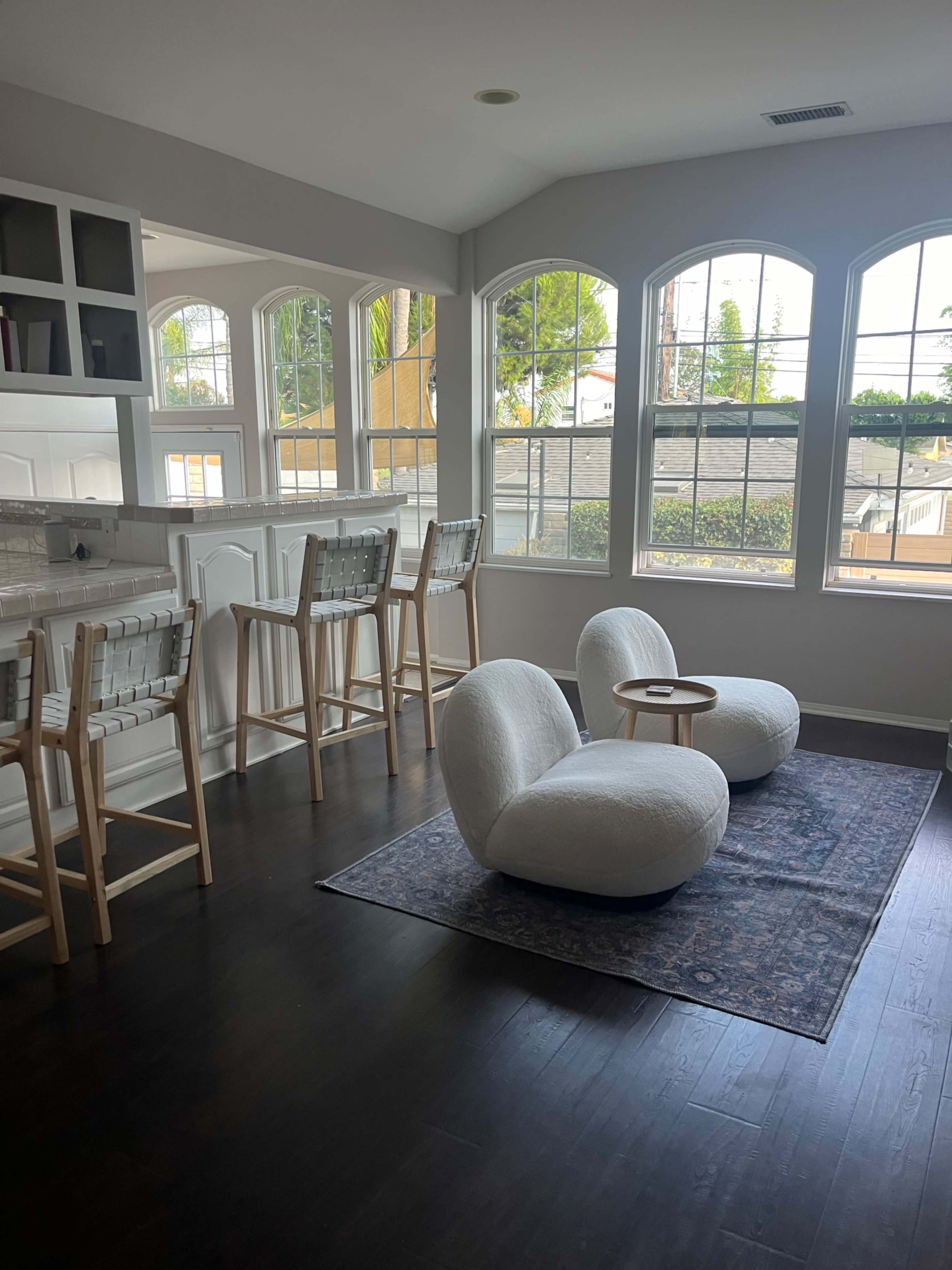 The image shows a modern, well-lit room featuring two soft chairs, a small side table, and a bar area with high stools, all positioned near large windows.