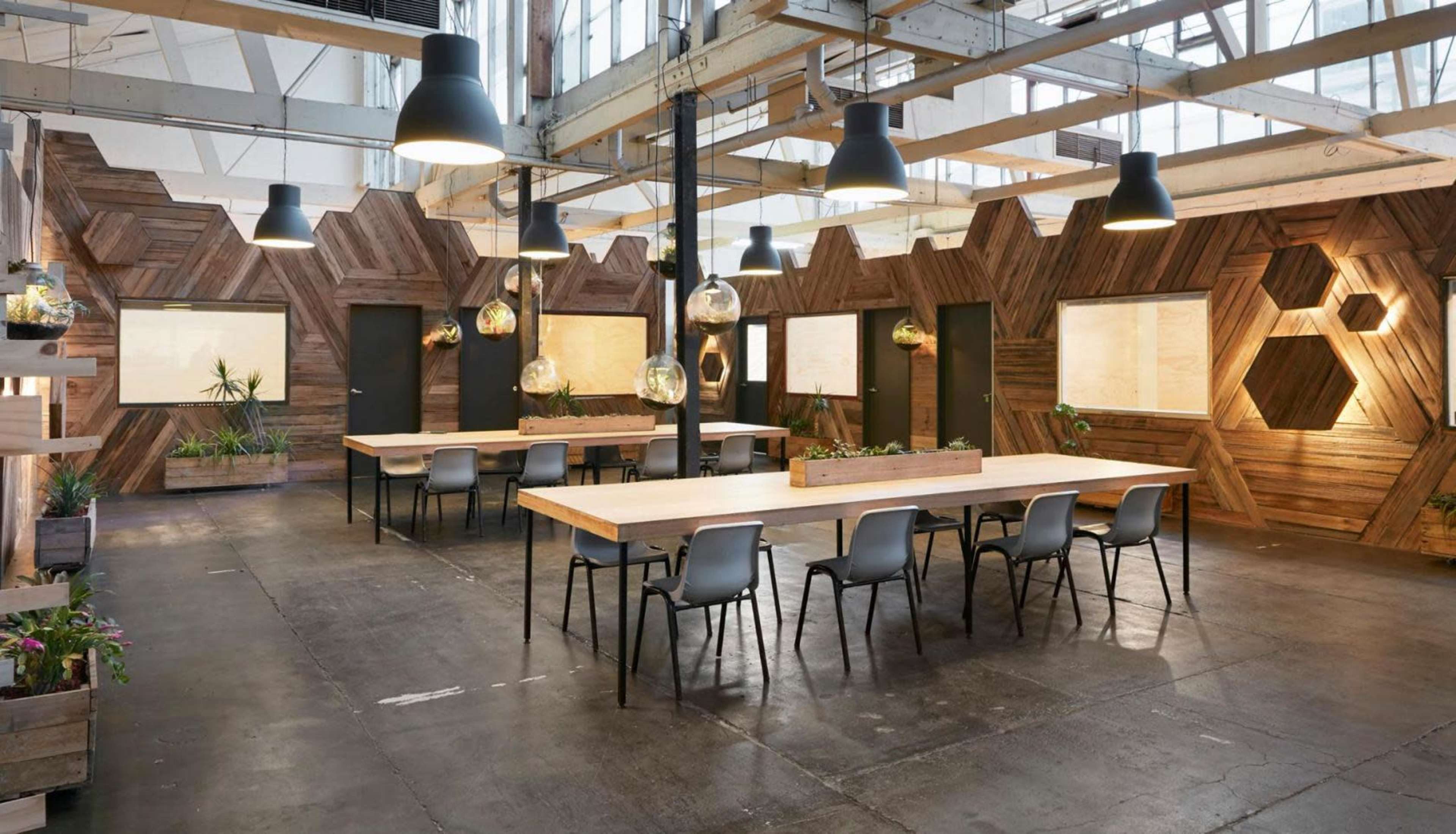 A modern, open workspace with wooden walls, large tables, black chairs, and pendant lighting, complemented by indoor plants.