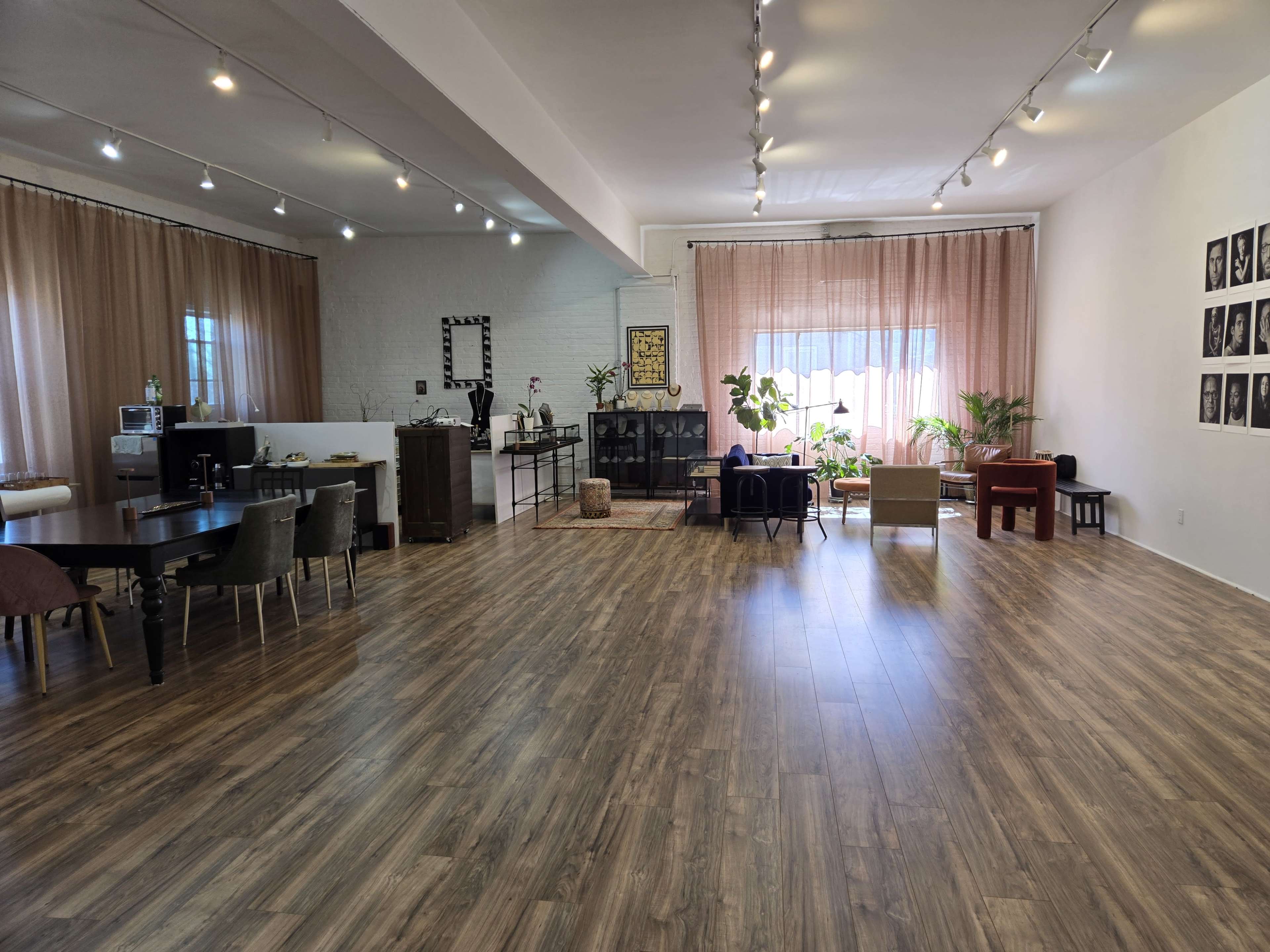 A spacious room with wooden flooring, equipped with a dining table, chairs, plants, and a large window draped with curtains.