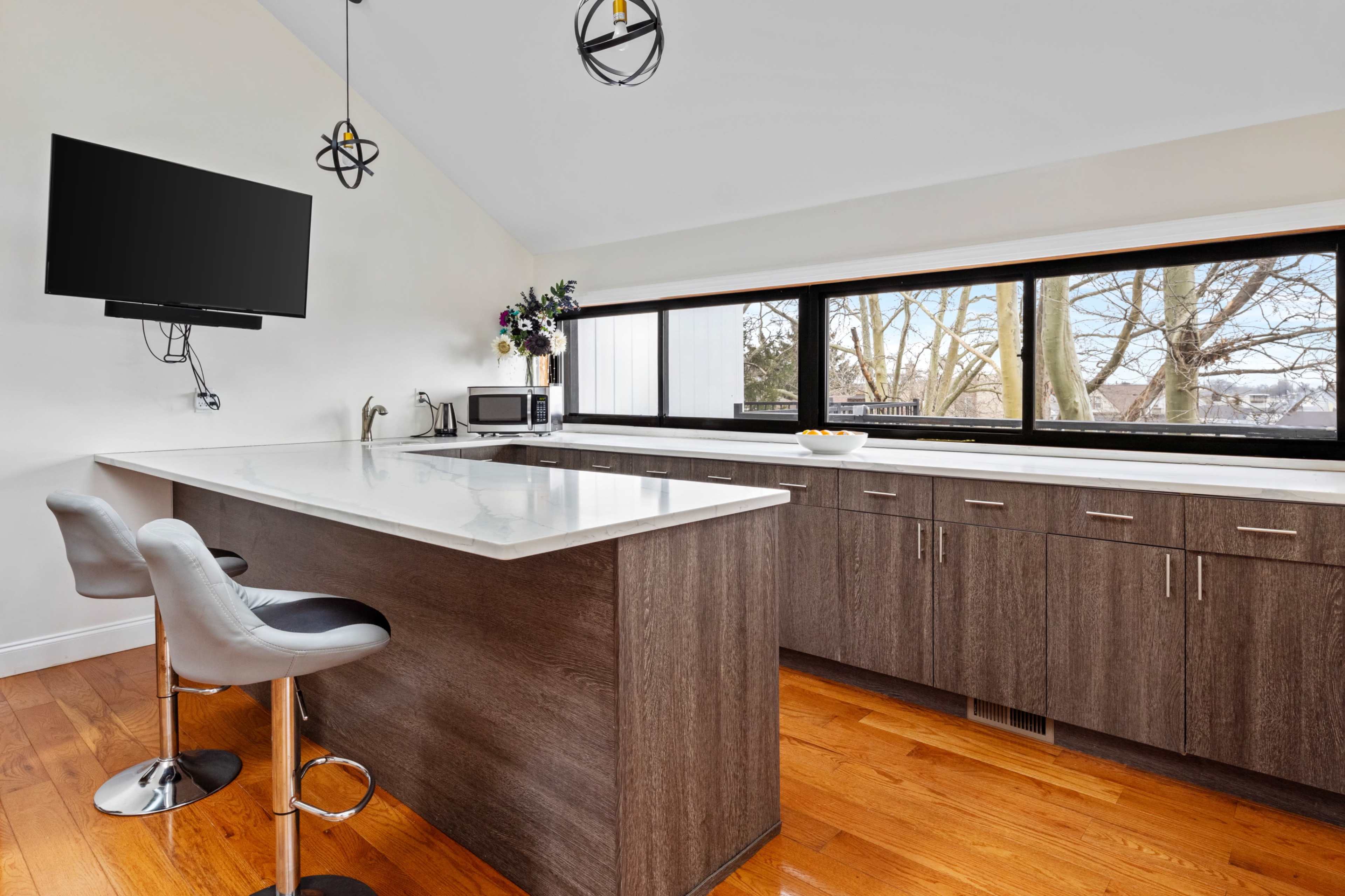 The kitchen features a modern design with a large island, wood cabinetry, and a mounted television beside a window.