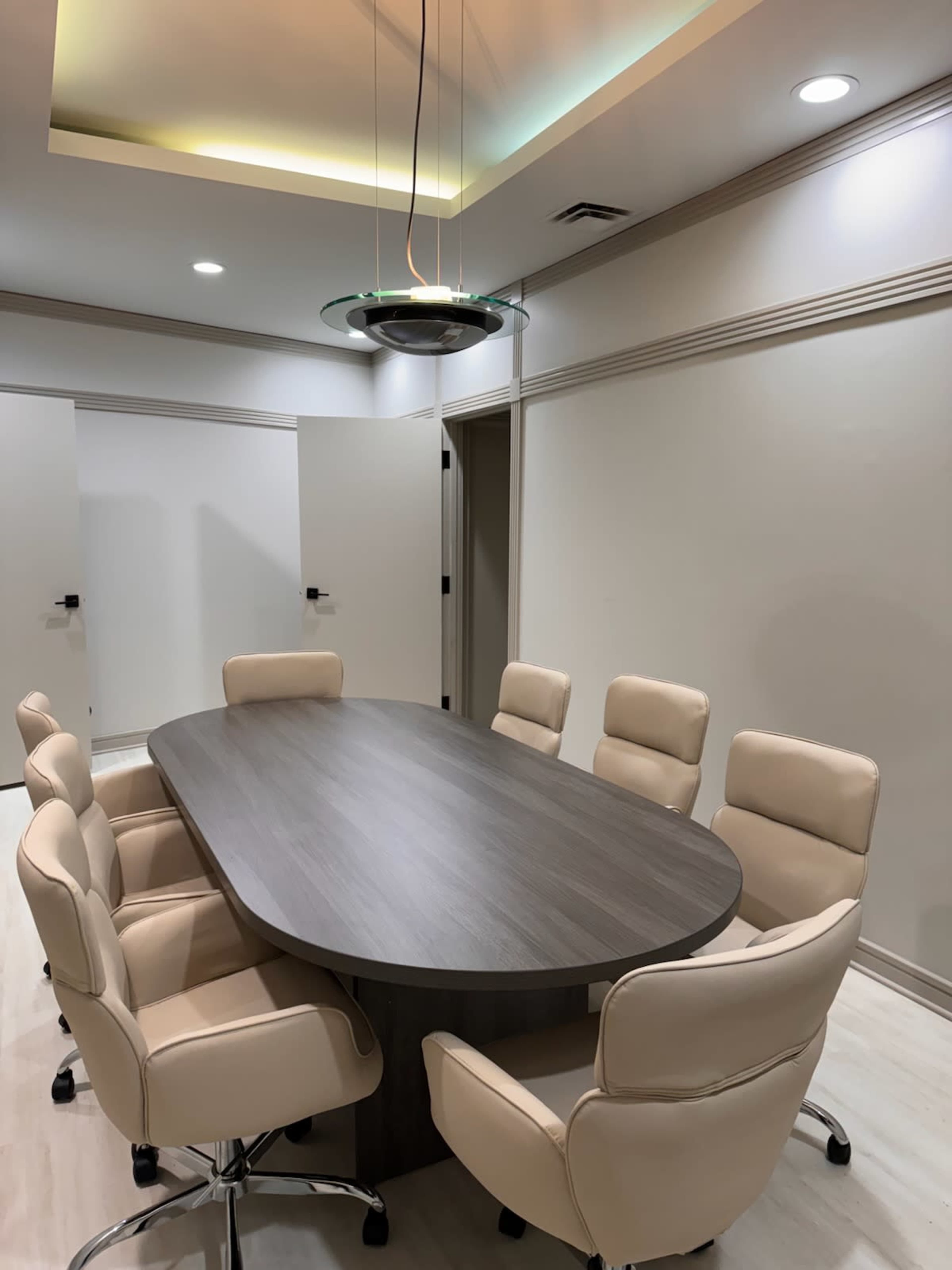 A modern conference room features an oval table surrounded by eight beige office chairs, with overhead lighting and neutral-colored walls.