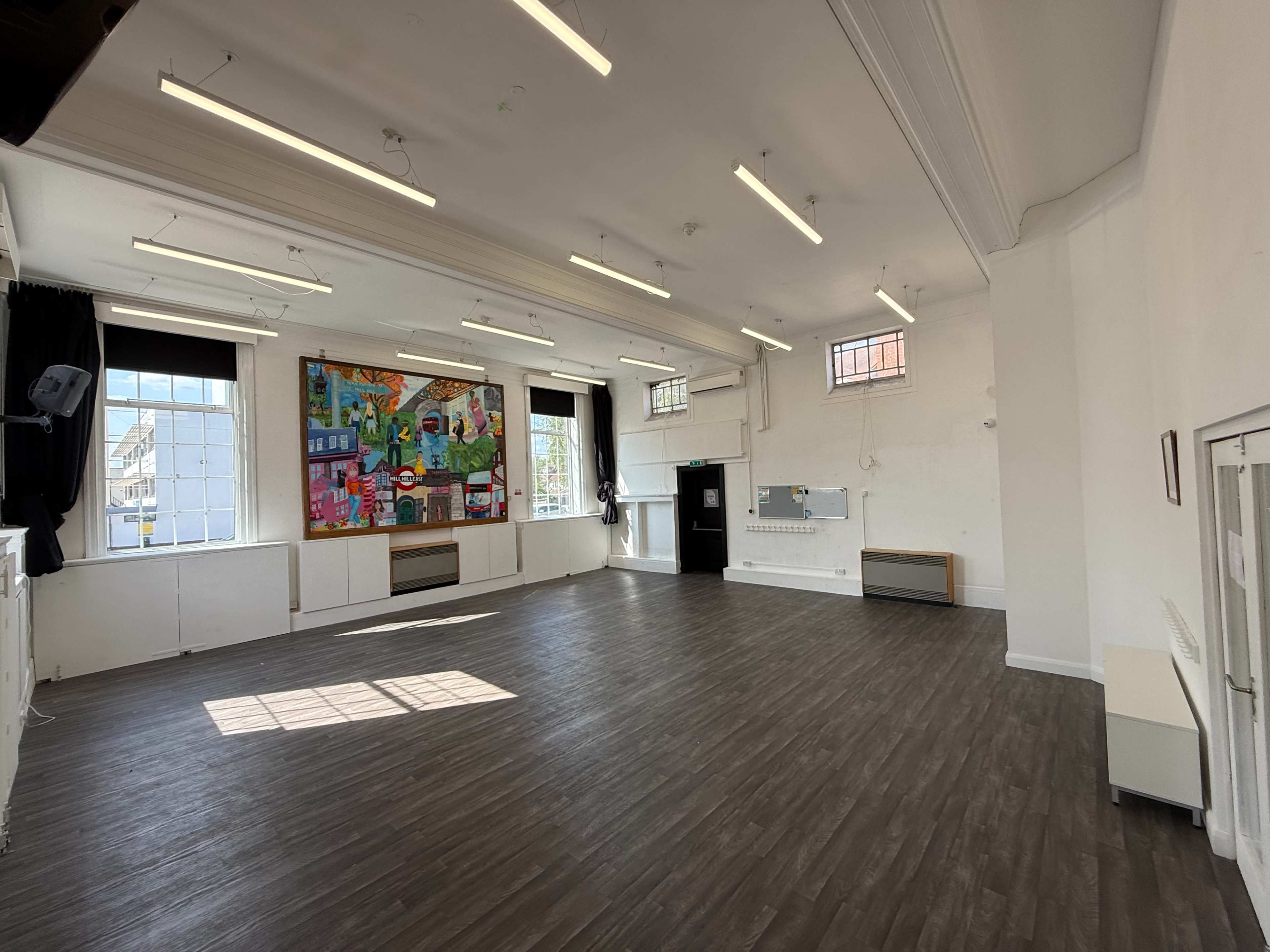 The image shows a spacious hall with wooden flooring, large windows, and a colorful mural on one wall.