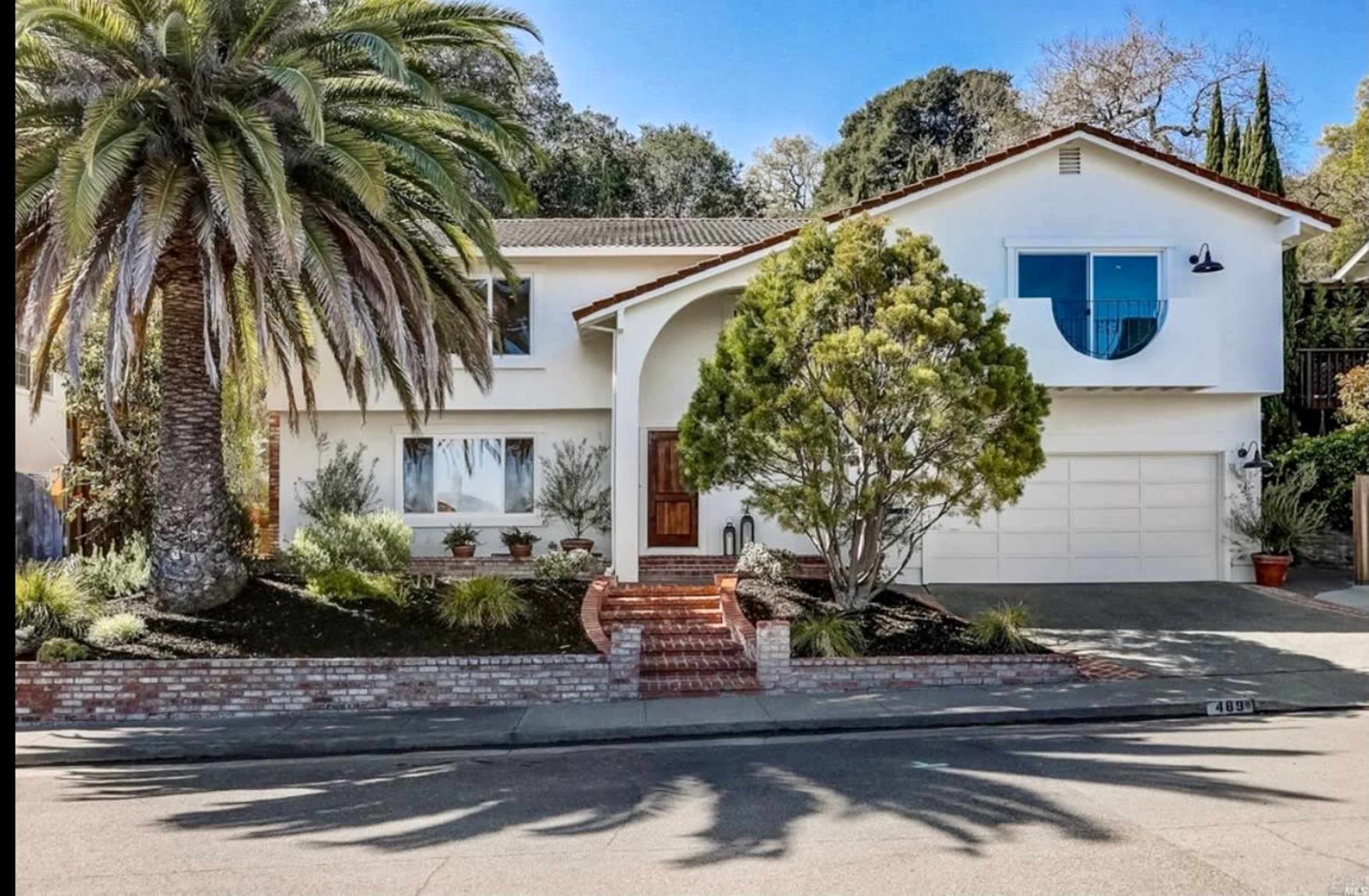 A two-story house with a palm tree and a circular window, situated on a sloped lot with a brick walkway leading to the entrance.