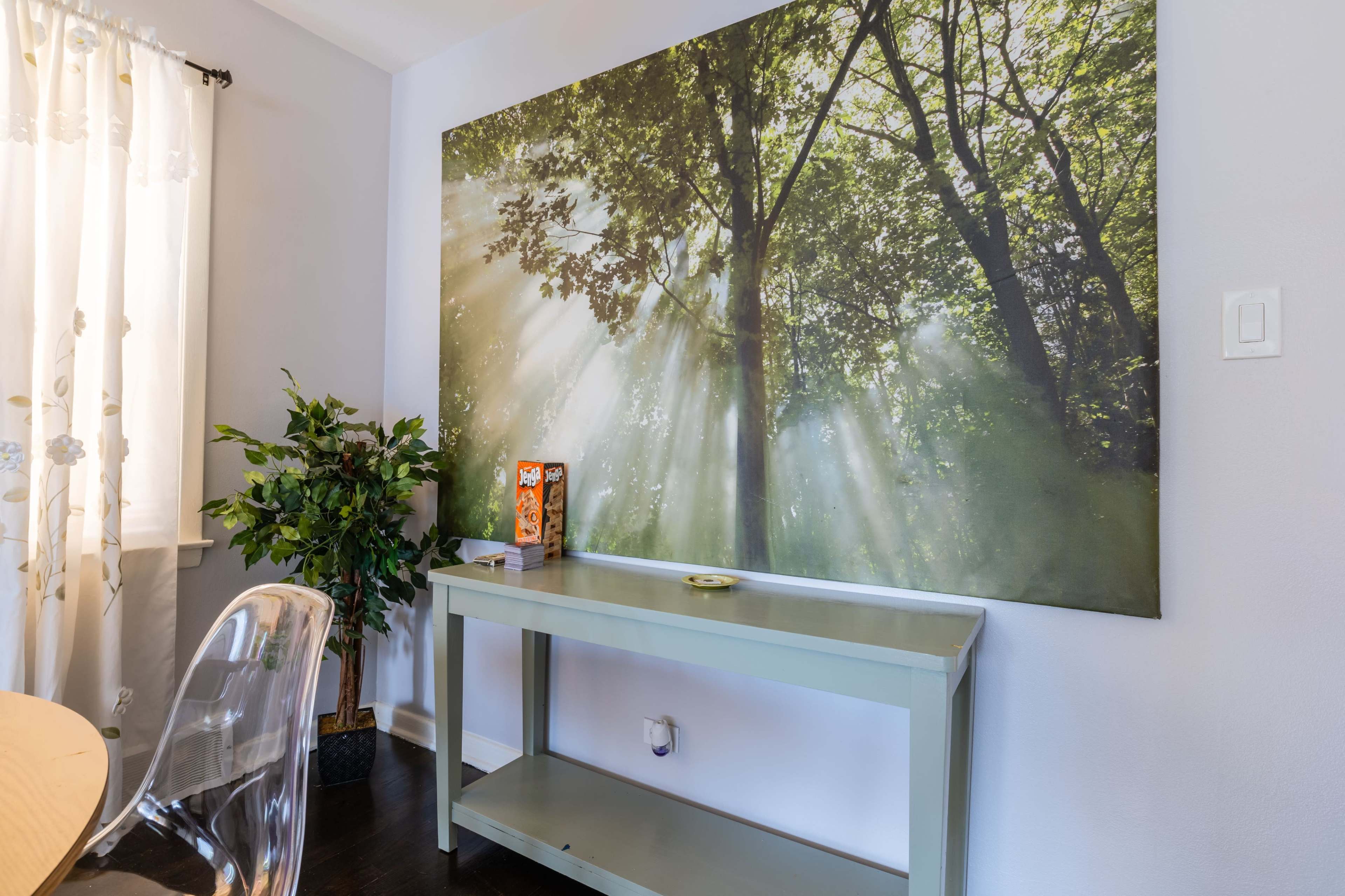 A light-filled room features a green table beneath a large wall mural of trees with sunlight filtering through their leaves.
