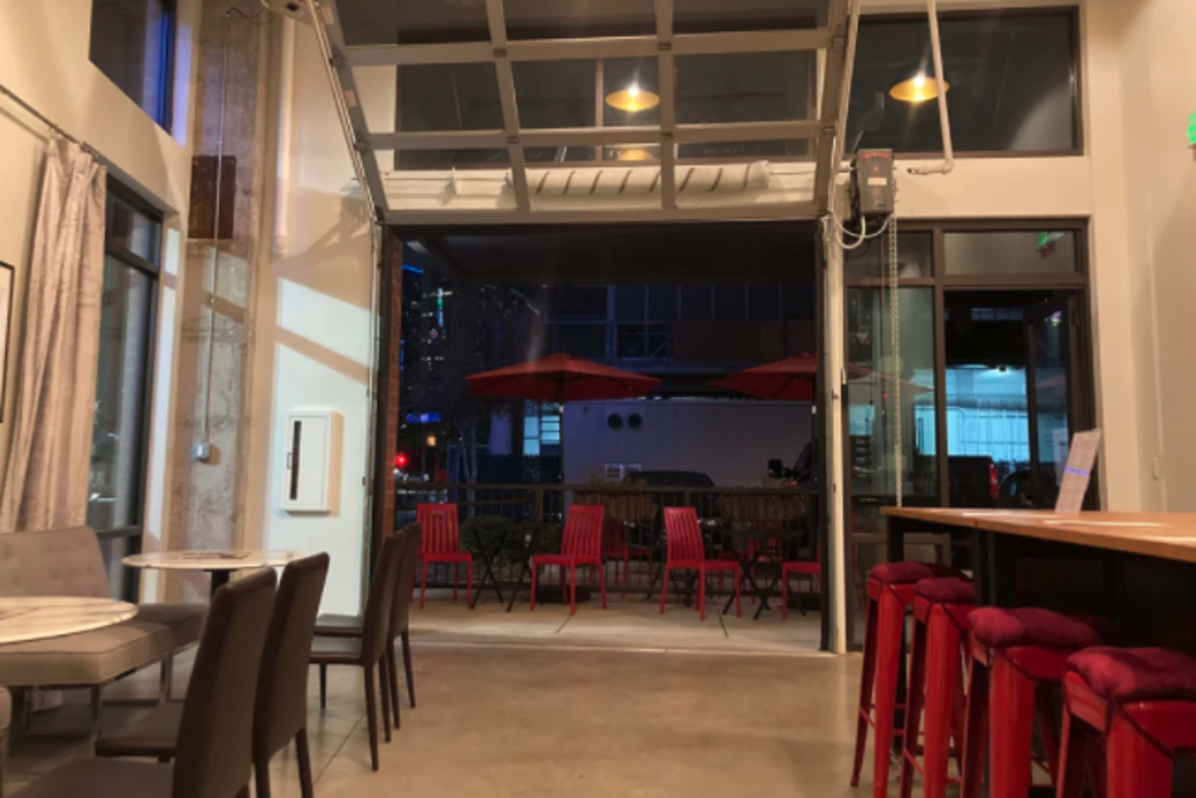 The interior of a modern café or bar, with a large open entrance leading to an outdoor seating area featuring red chairs and umbrellas.