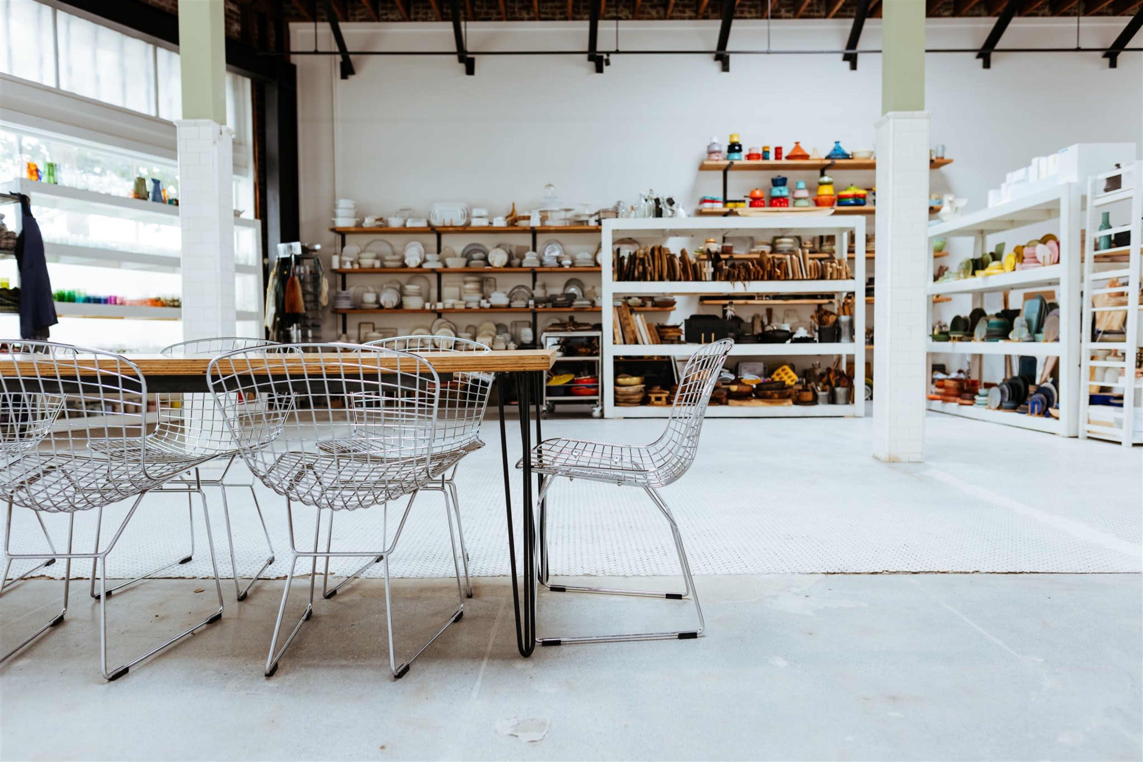 A spacious interior features a wooden table surrounded by wire chairs, with shelves filled with colorful kitchenware in the background.