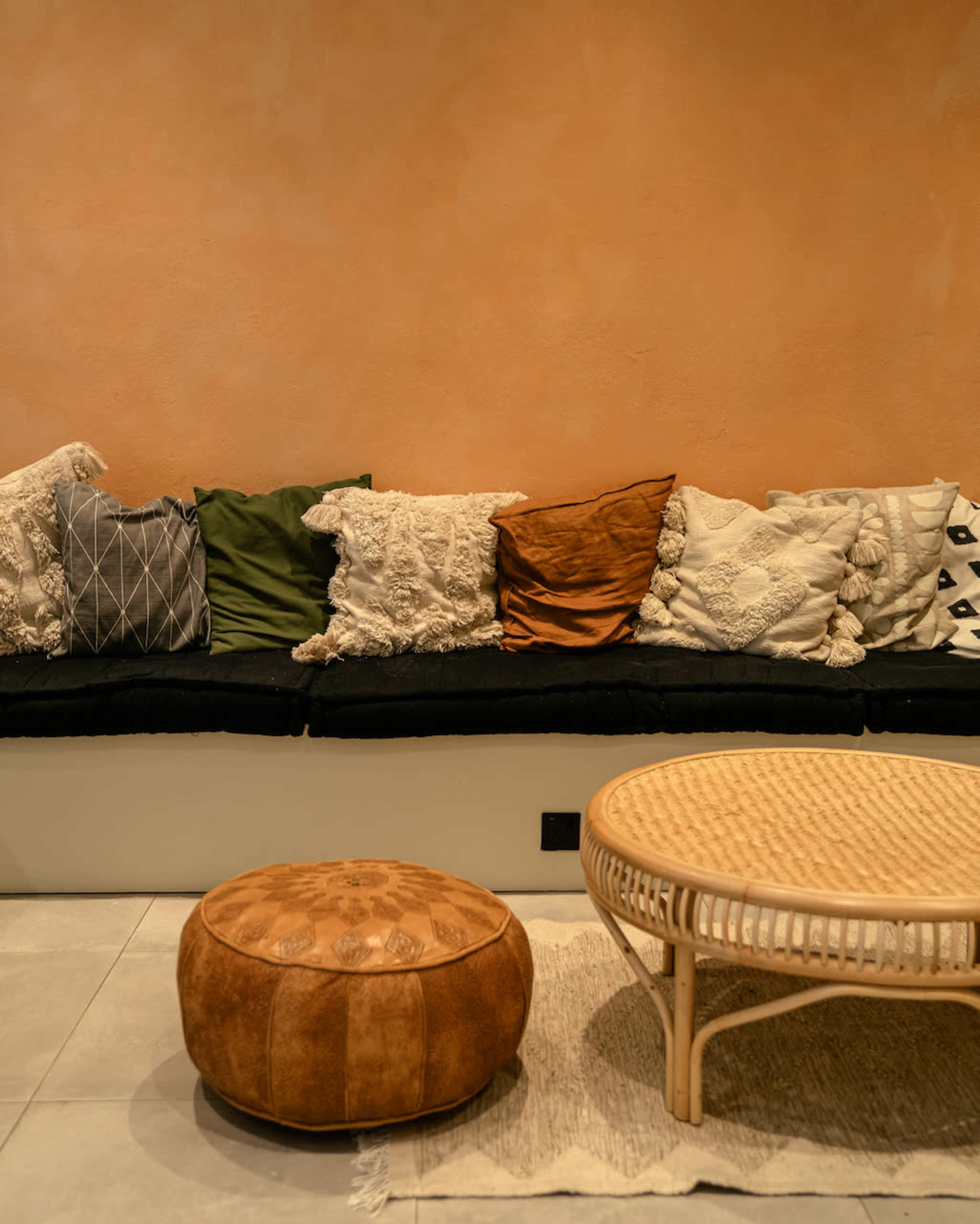 A cozy seating area with a black cushioned bench adorned with various patterned pillows and a round rattan coffee table next to a leather pouf.