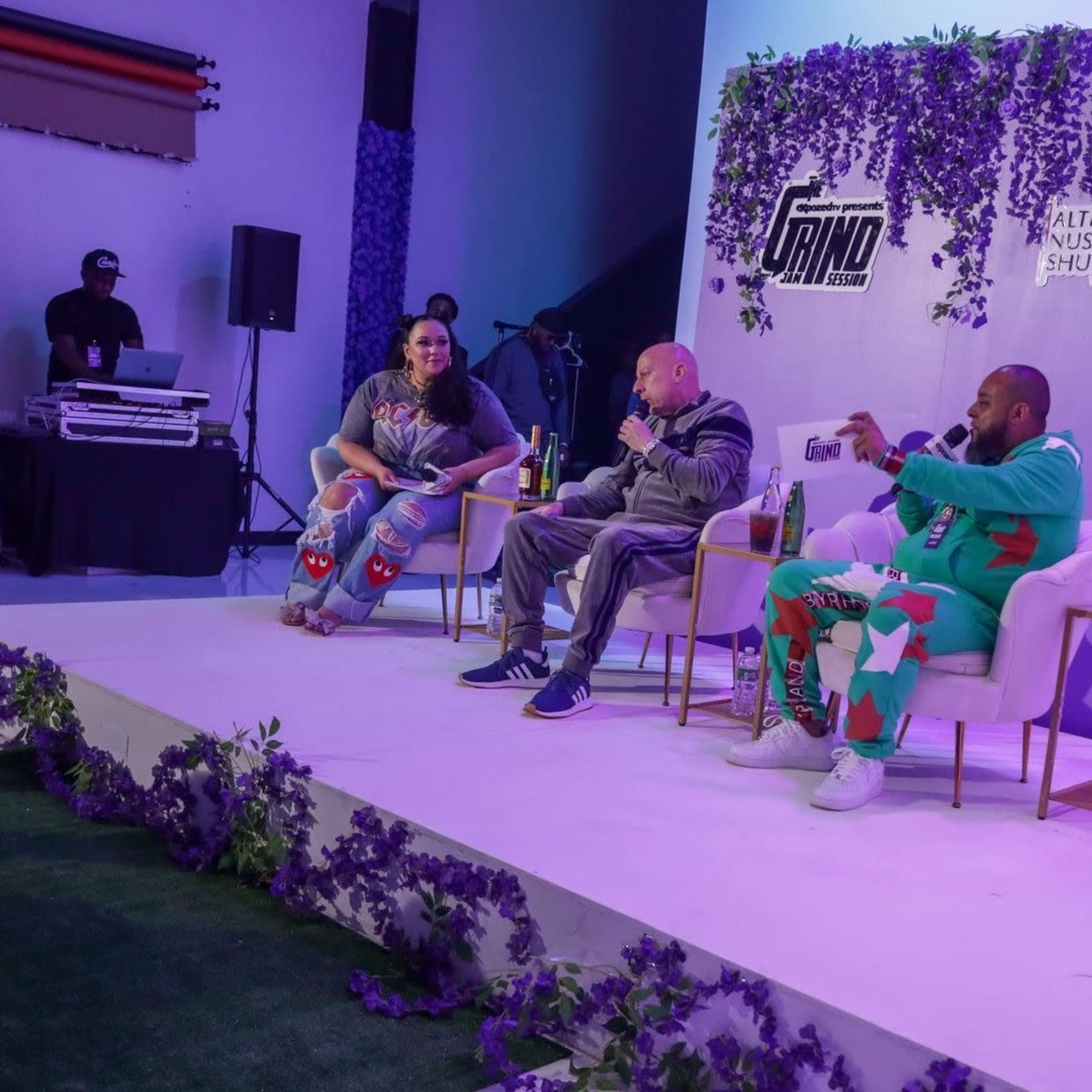 A panel discussion takes place on stage, with three individuals seated and a DJ setup visible in the background.