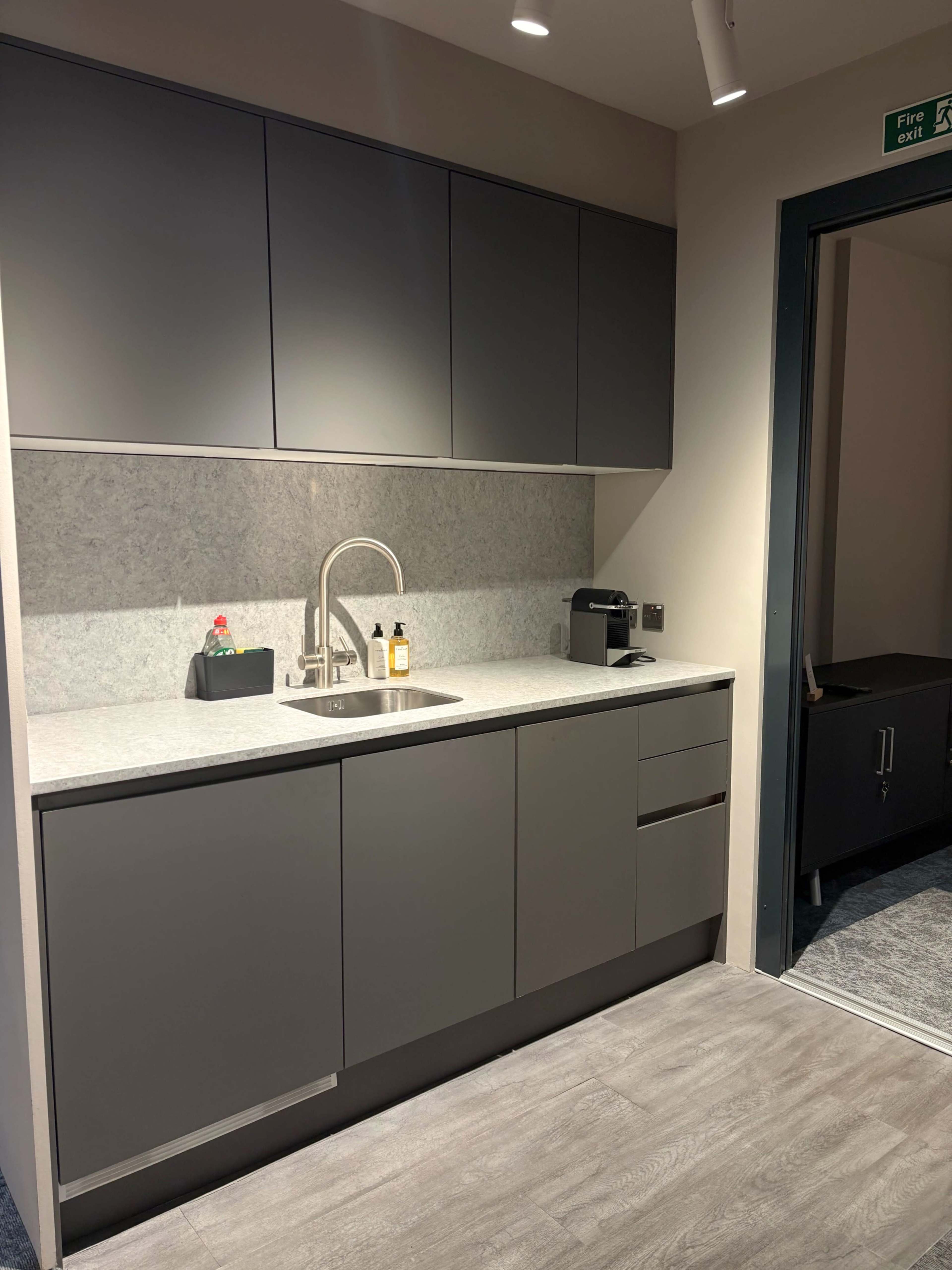 The image depicts a modern kitchen area with gray cabinets, a stainless steel sink, a countertop, and a doorway leading to another space.