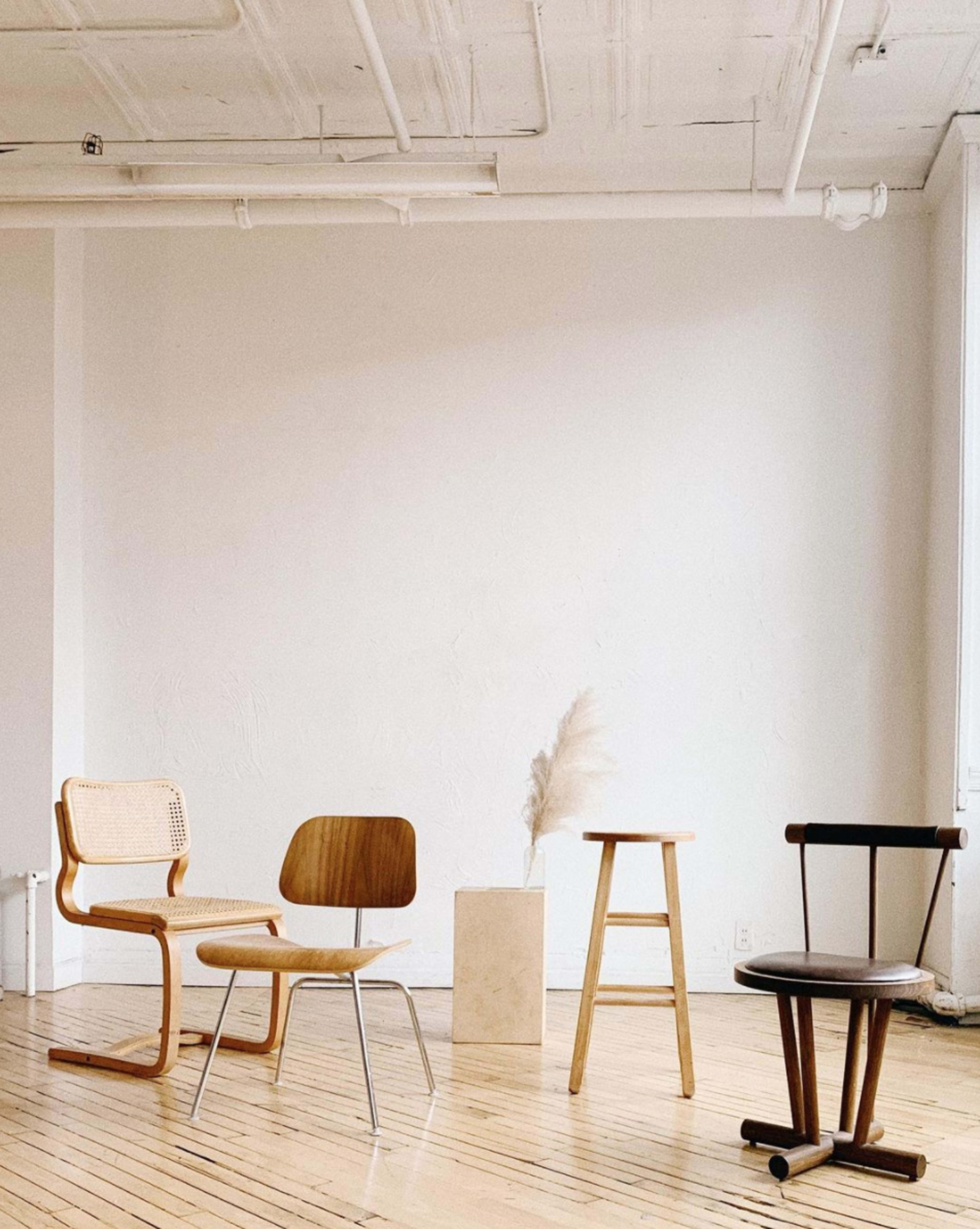 Four different styles of chairs arranged in a minimalist space with a light-colored wall and a simple pedestal displaying a decorative plant.