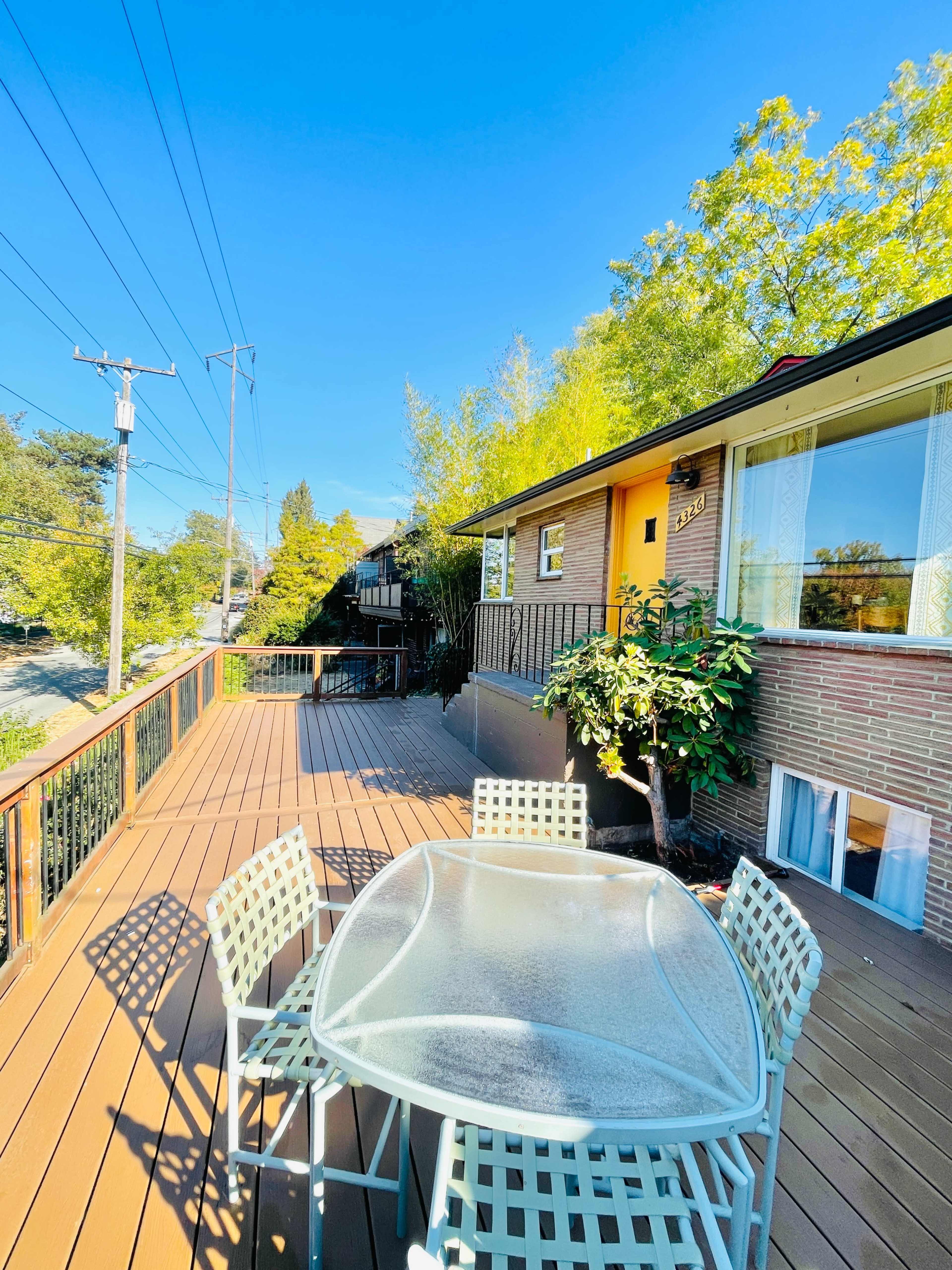 A house with a wooden deck features a glass table and chairs, surrounded by greenery under a clear blue sky.