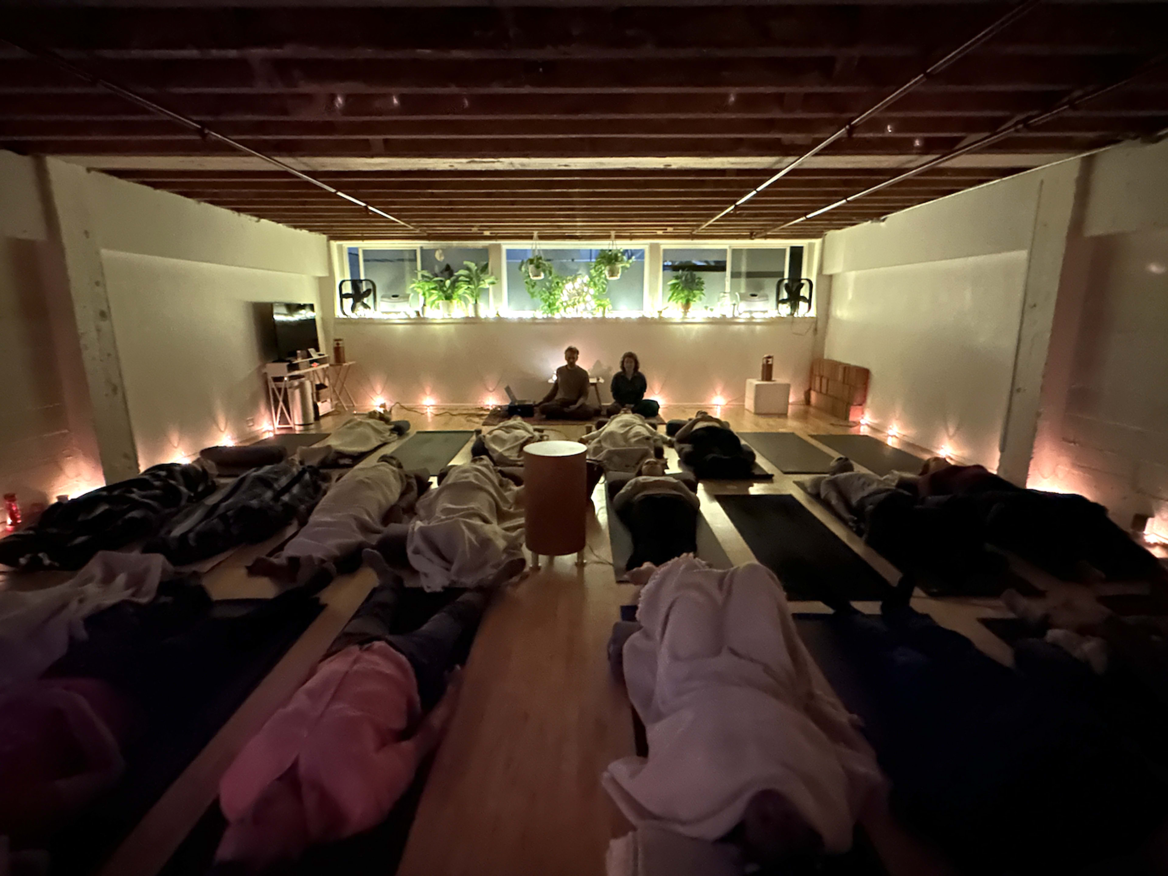 A yoga studio with participants lying on mats in a dimly lit room surrounded by candles and plants.