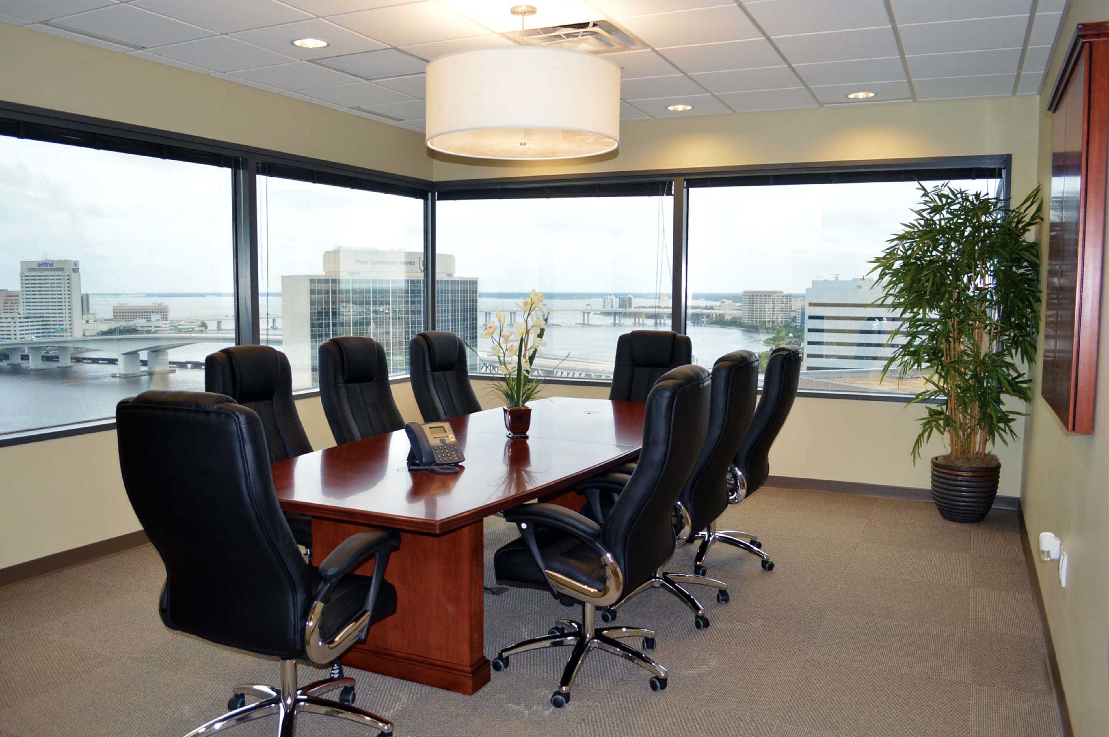 A large conference room features a wooden table surrounded by black swivel chairs, with windows overlooking a view of a river and city skyline.