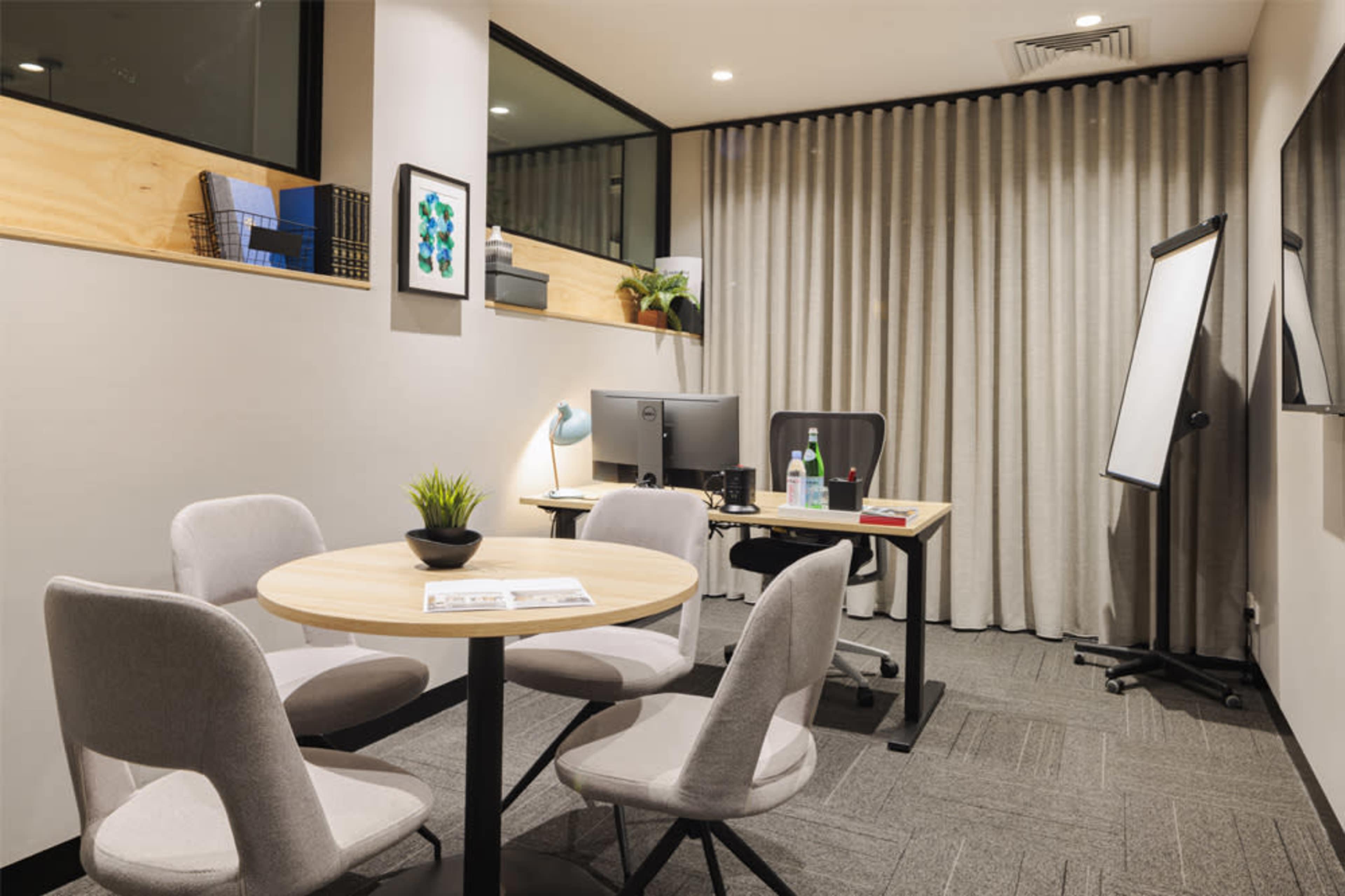 A modern office space featuring a round table with four chairs, a computer desk with a monitor, and a presentation board against a backdrop of curtains.