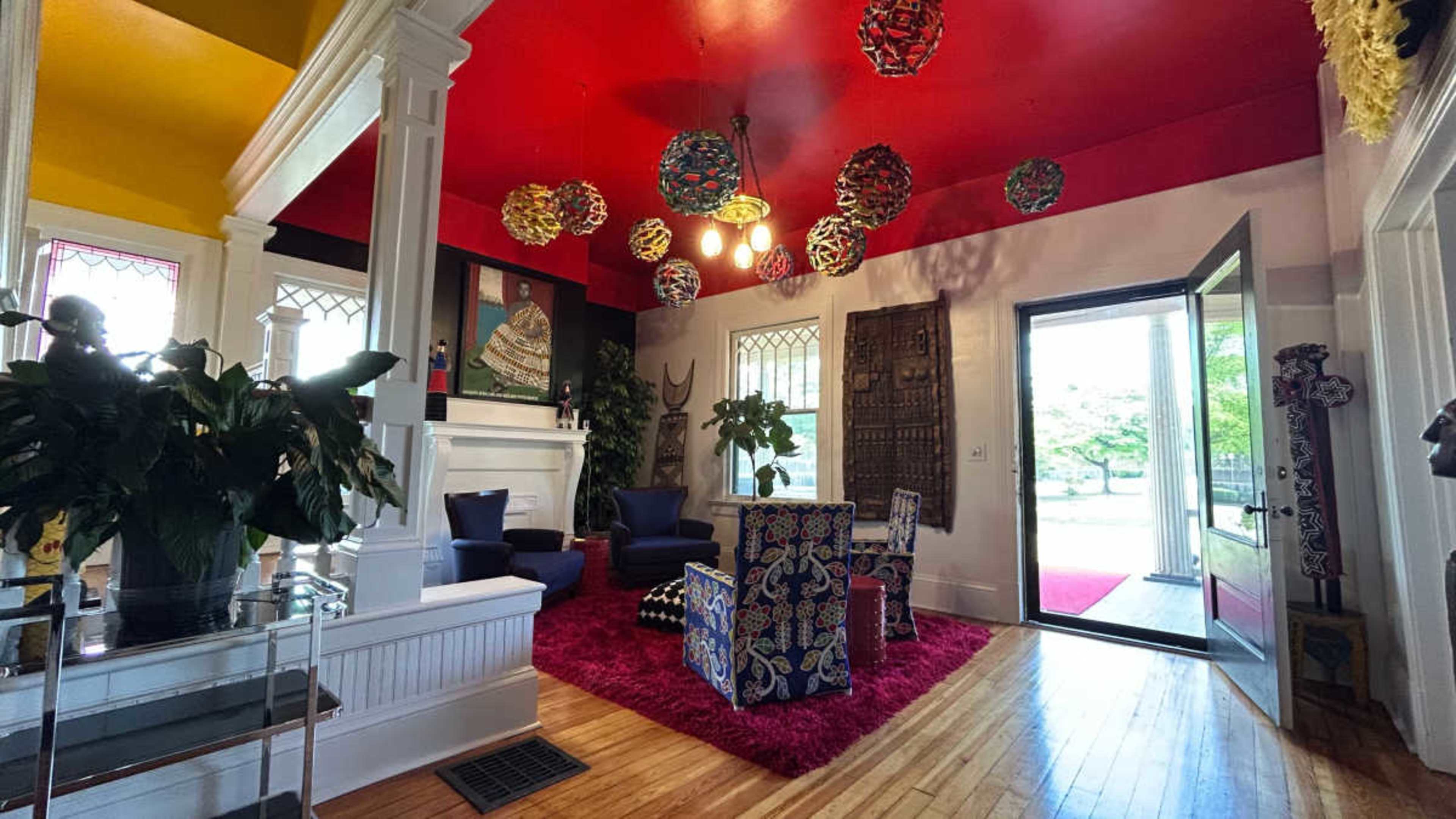The scene shows a vibrant entryway featuring a red and yellow ceiling, colorful hanging decorations, and two armchairs beside a doorway leading to an outdoor view.