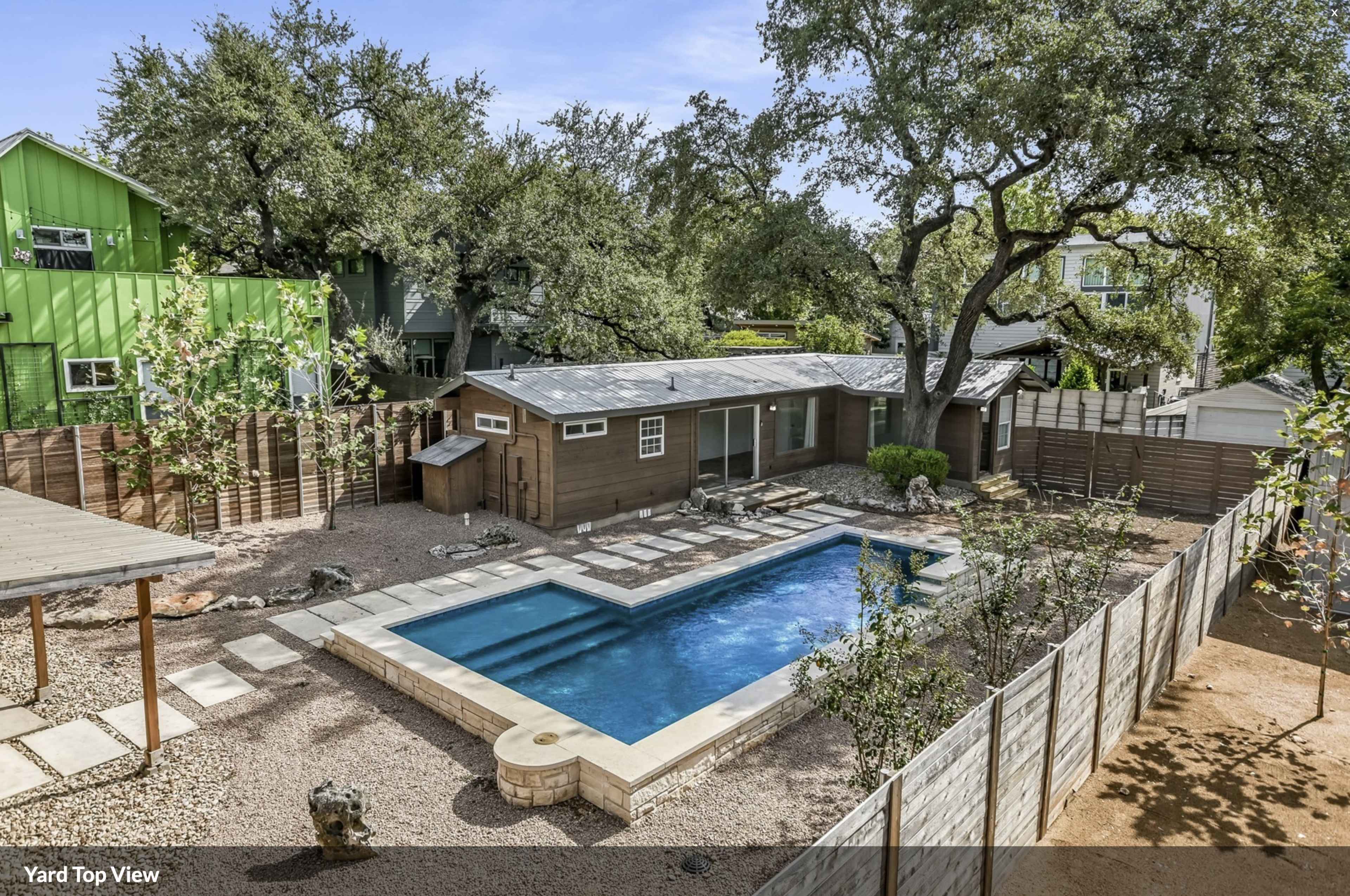 The image shows a backyard featuring a swimming pool, a wooden deck, landscaping with stones, and surrounding trees and houses.