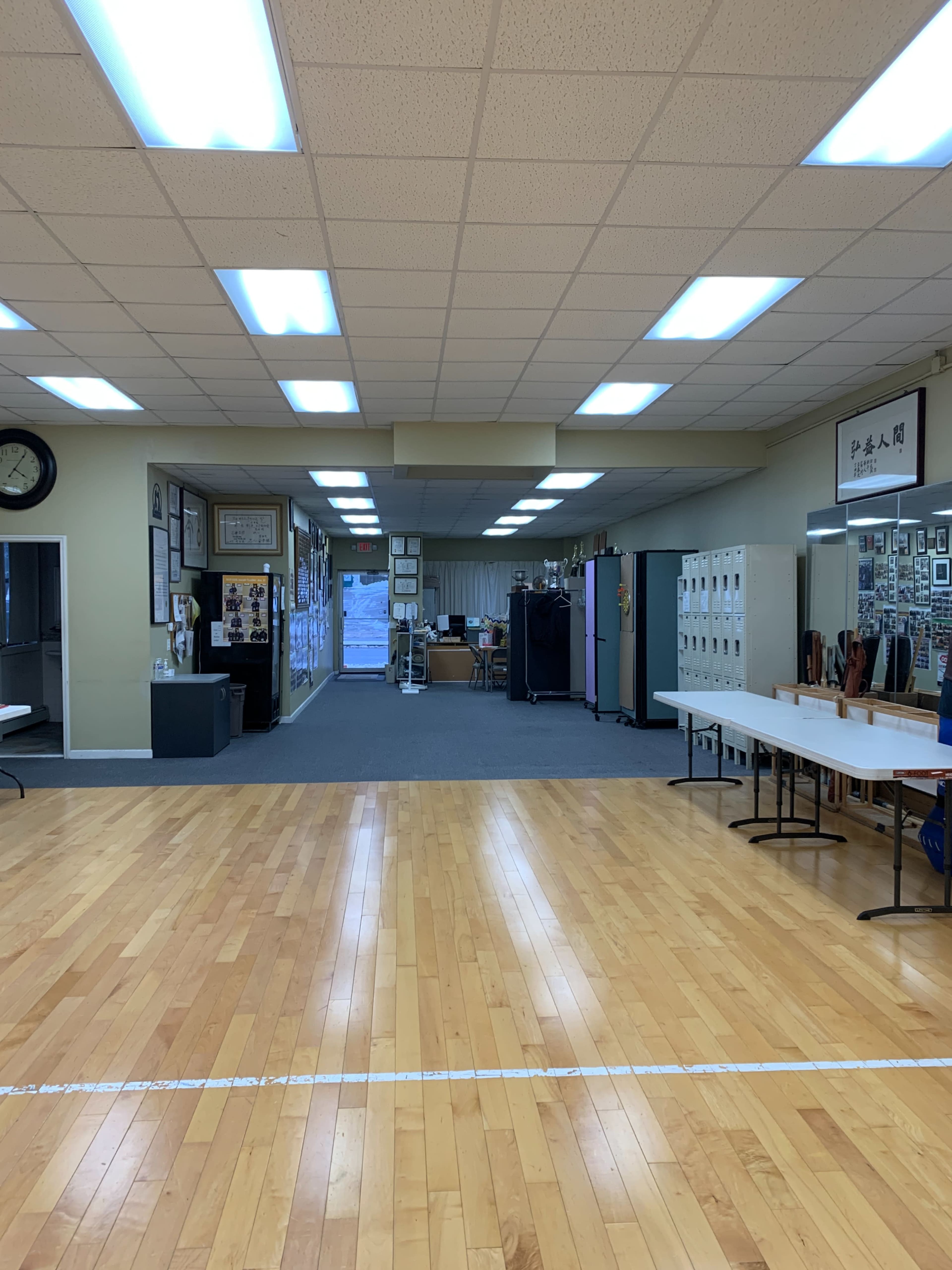 The image shows a large, empty indoor space with wooden flooring, leading to a room with lockers and various wall decorations.