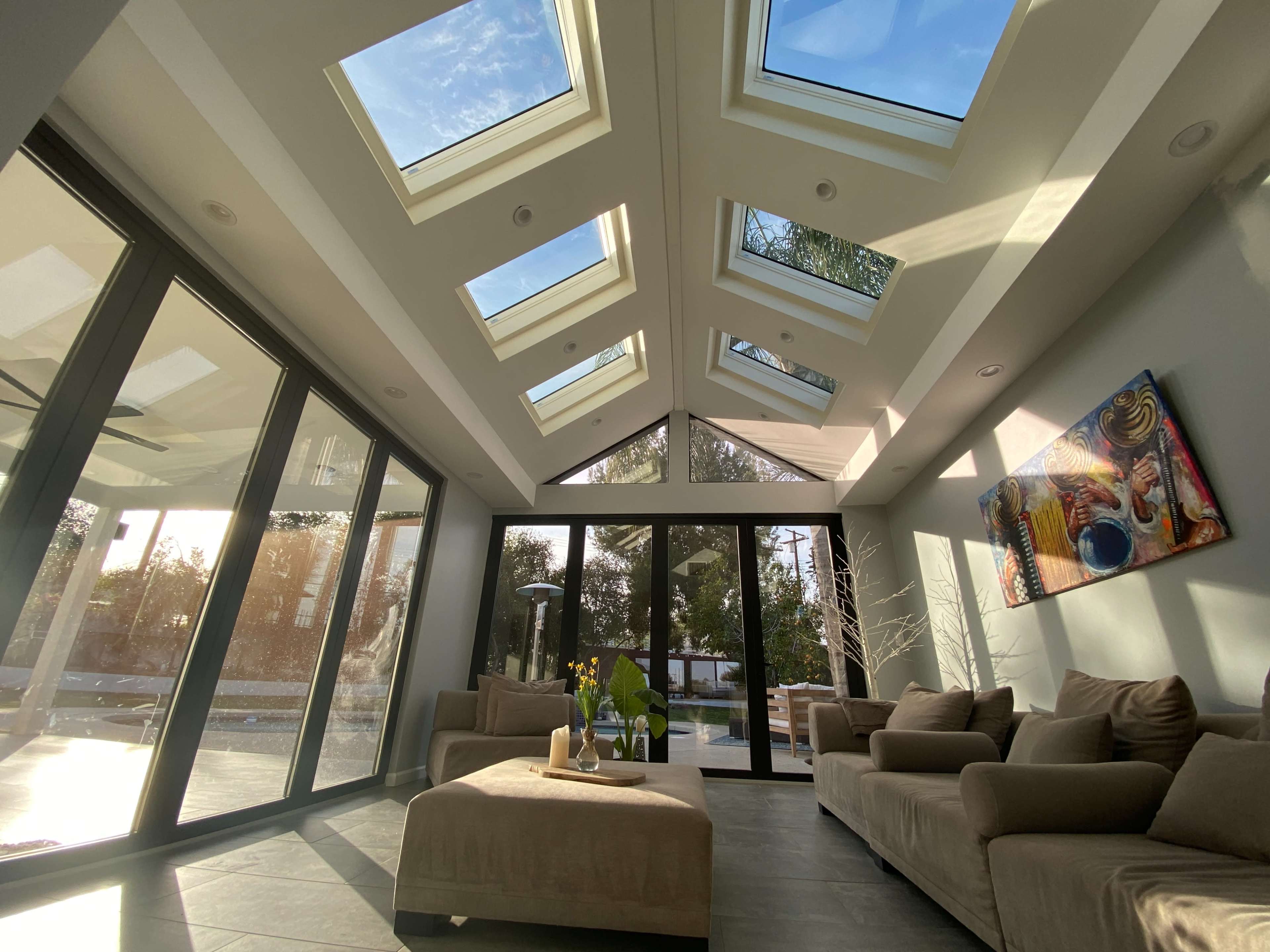 A sunlit modern living room with large skylights and glass walls, featuring beige sofas and a decorative painting on the wall.
