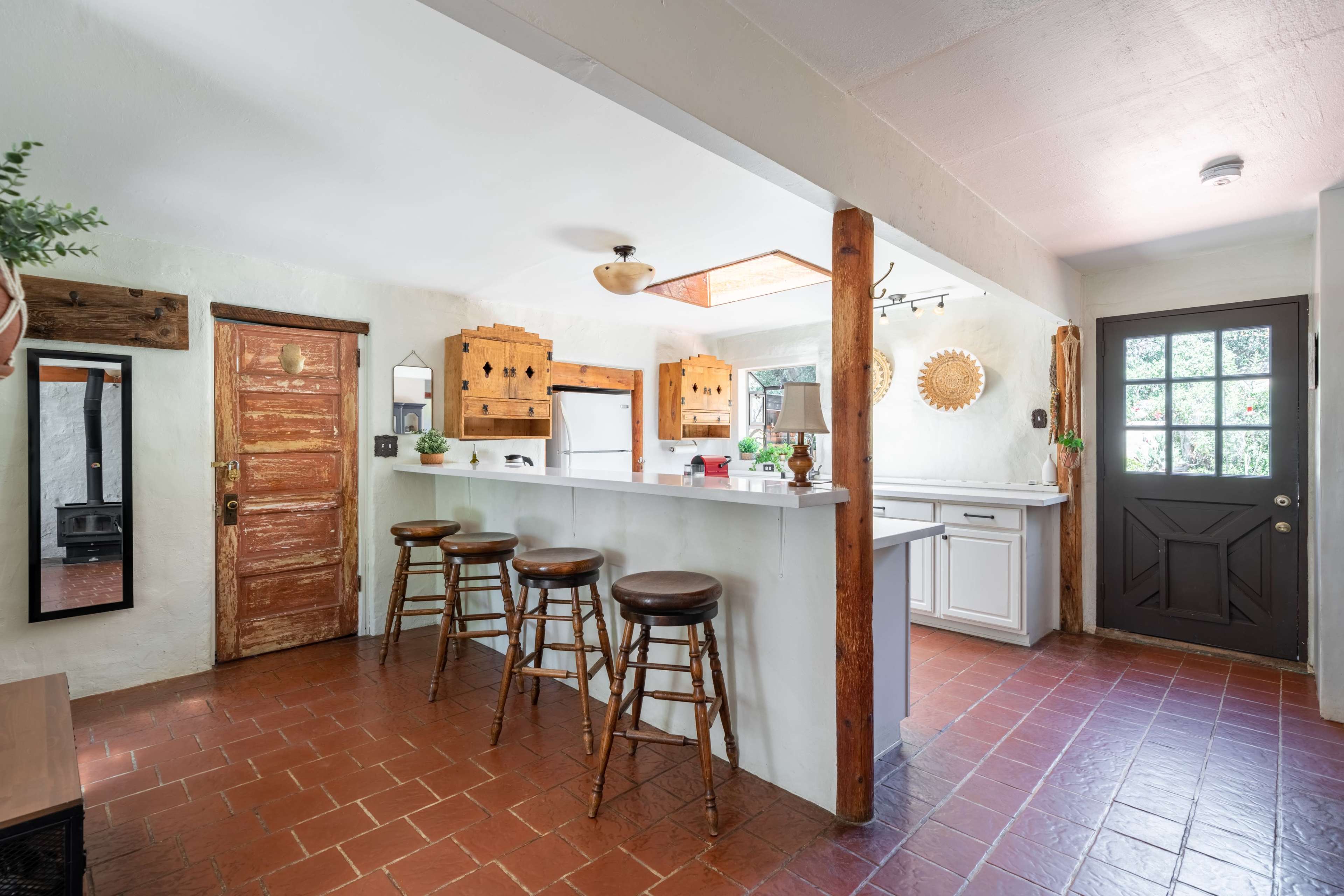 A kitchen with a bar area featuring three wooden stools, a wooden door, and natural light coming in from a window.