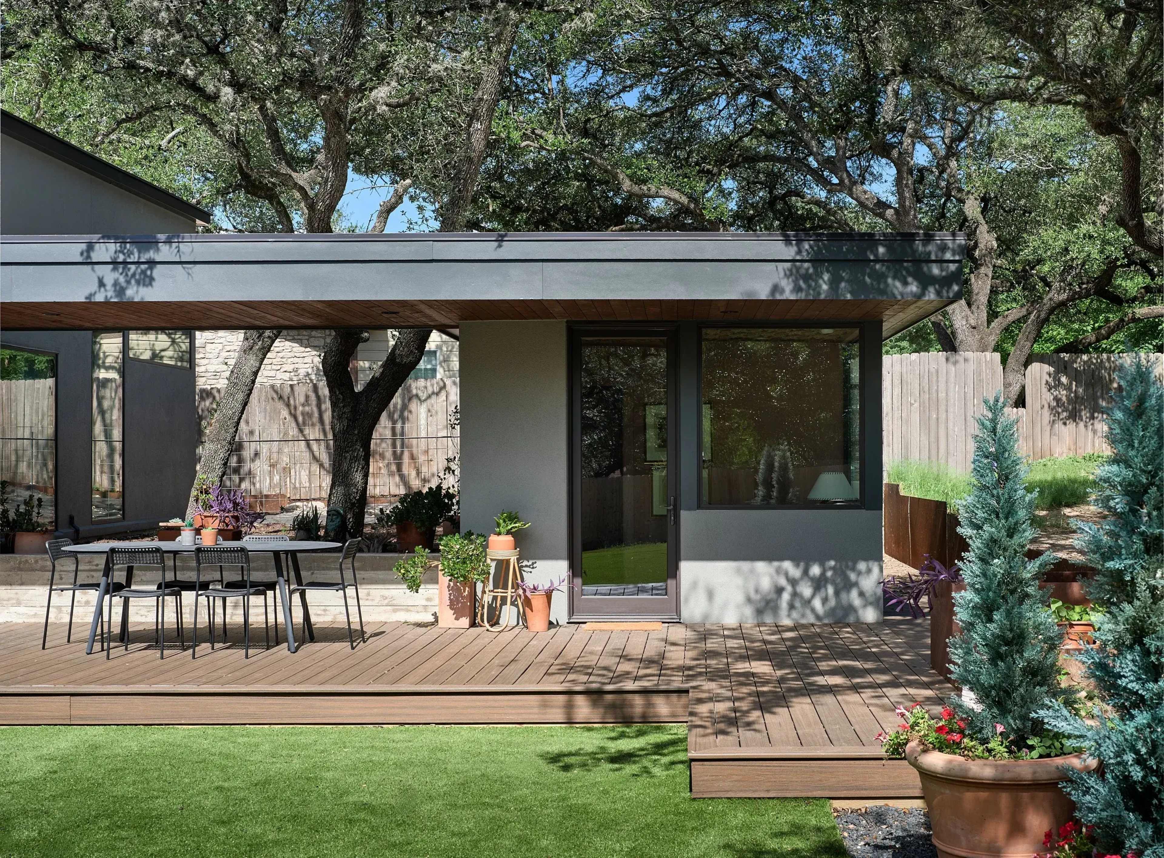 The image shows a modern outdoor patio area with a wooden deck, a dining table, and various potted plants surrounded by trees and a fence.