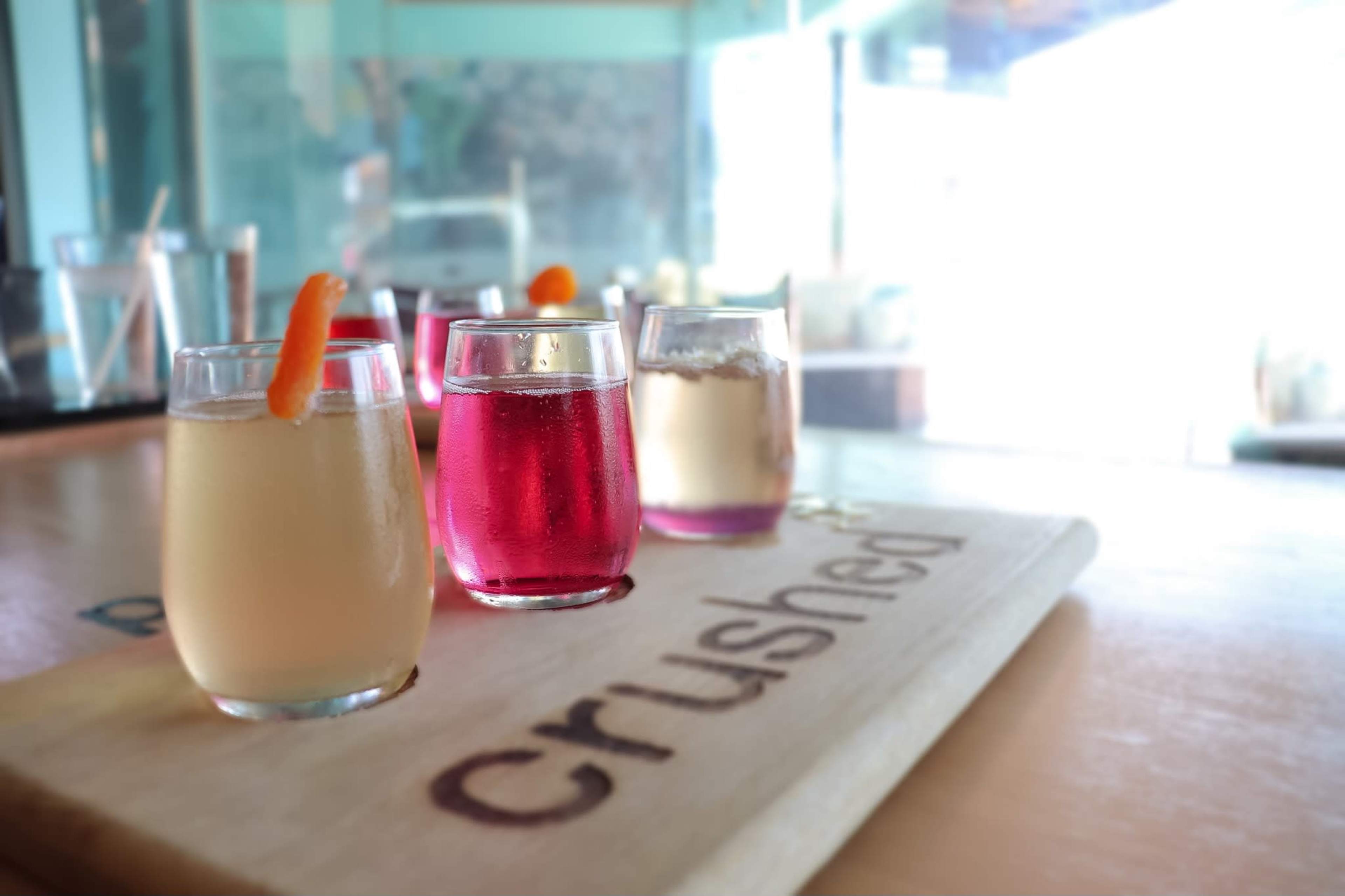 Three small glasses of colorful drinks arranged on a wooden tray labeled "crushed."