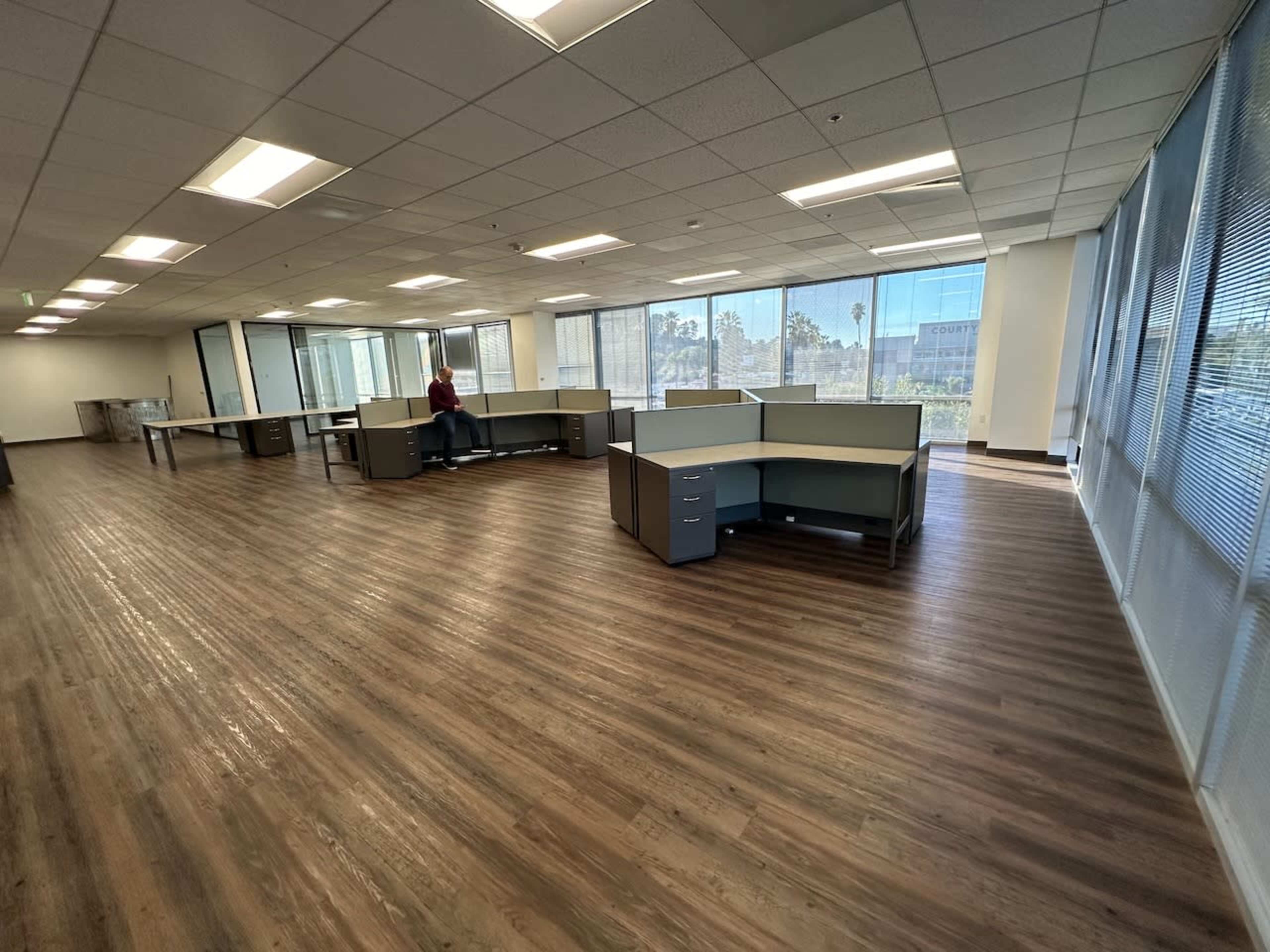 An office interior features several cubicles arranged in an open space with large windows allowing natural light.