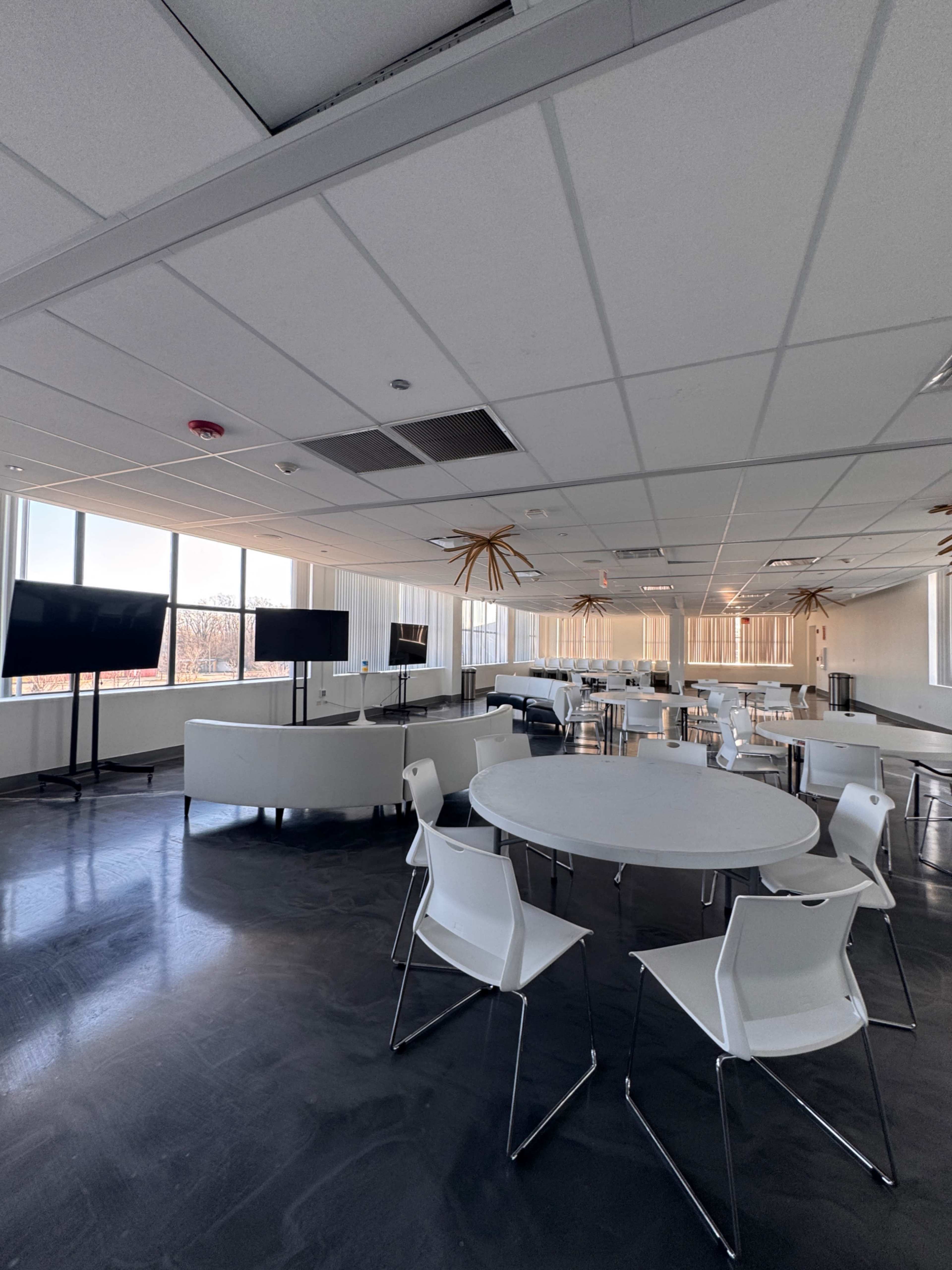 The image shows a modern office space with white furniture, including tables and chairs, alongside multiple television screens and large windows allowing natural light.