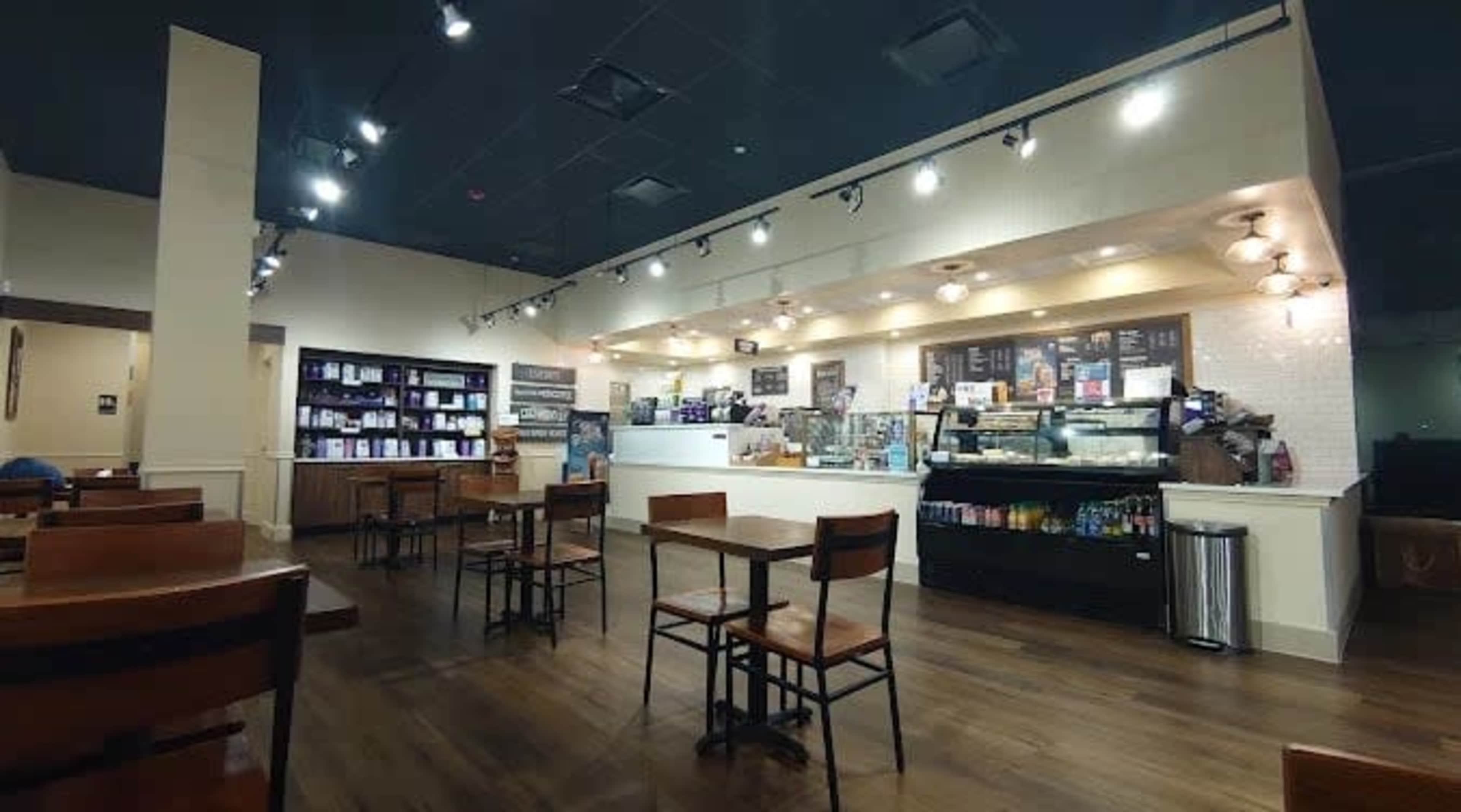 The image shows a spacious coffee shop interior with several wooden tables and a counter displaying various food and beverage options.