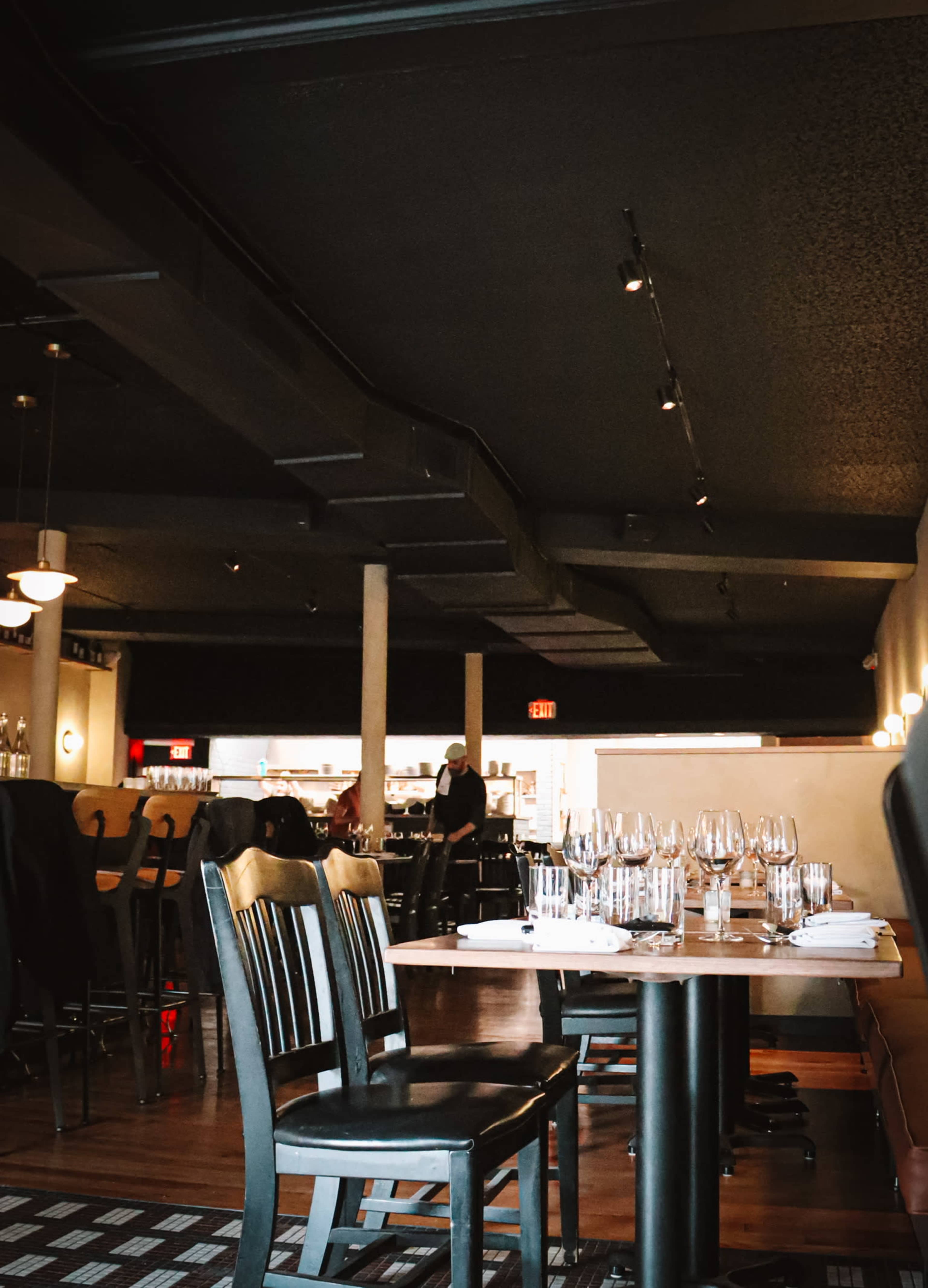 The image shows an empty restaurant interior with wooden chairs and tables set for dining, and a server moving in the background.