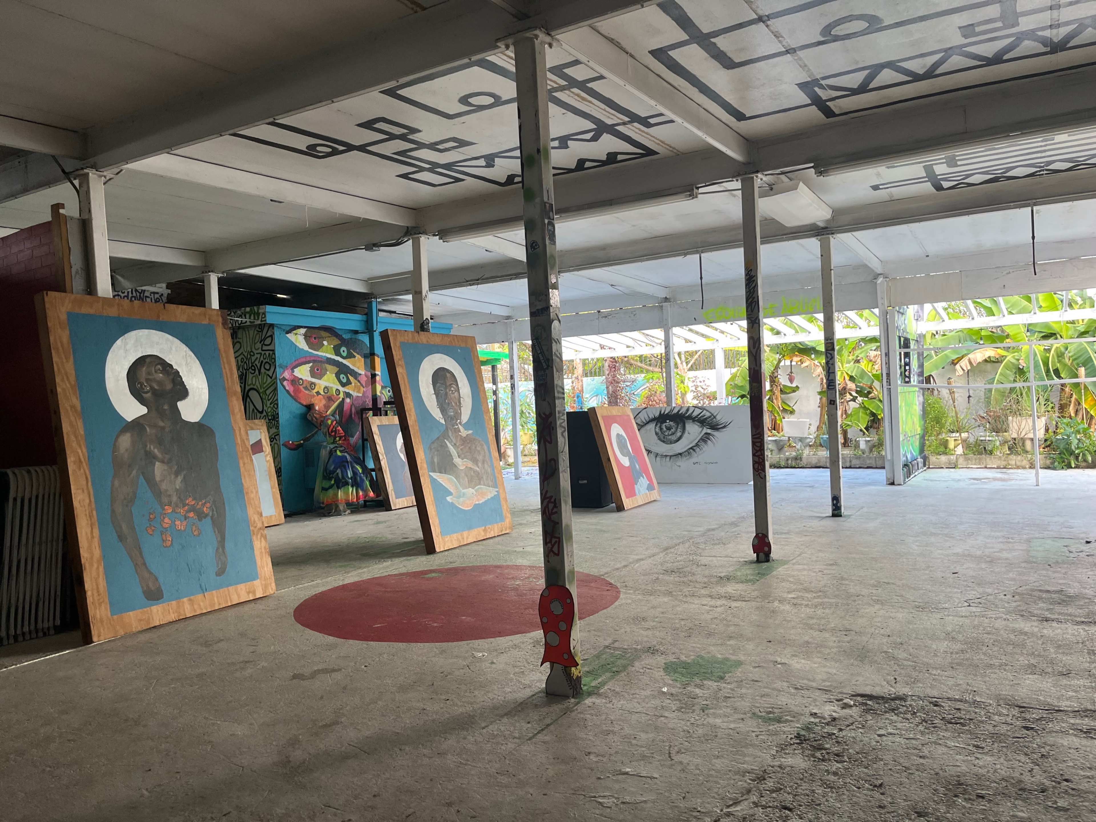 An indoor space featuring large, colorful artwork displayed against a backdrop of vibrant murals and greenery visible through open doors.