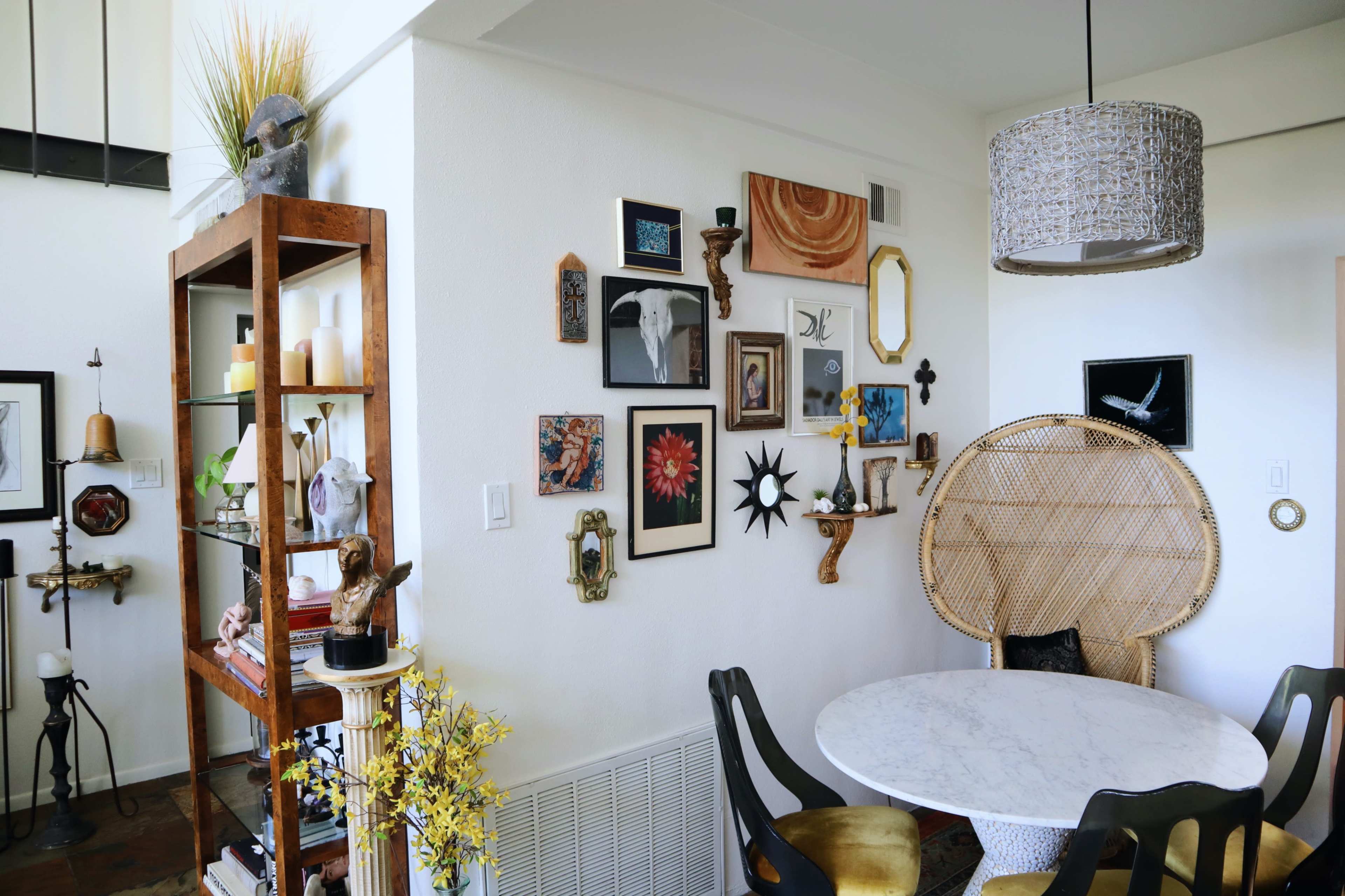 A stylish room with a round marble table, a woven chair, and a wall decorated with various framed artworks and decorative objects, along with a bookshelf filled with small items and plants.