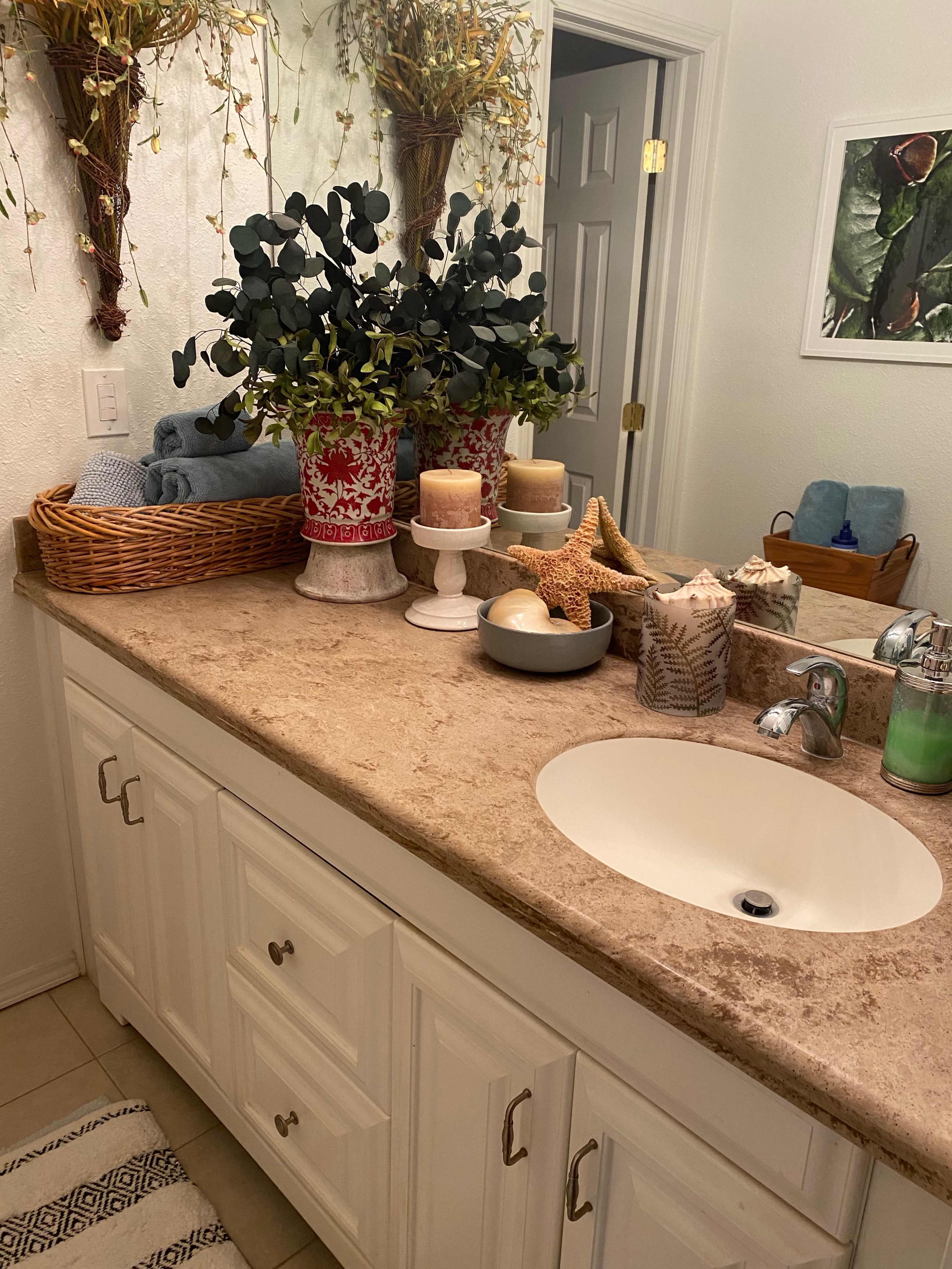The image shows a neatly arranged bathroom countertop featuring decorative plants, candles, and a basket, with a sink and white cabinetry in the background.