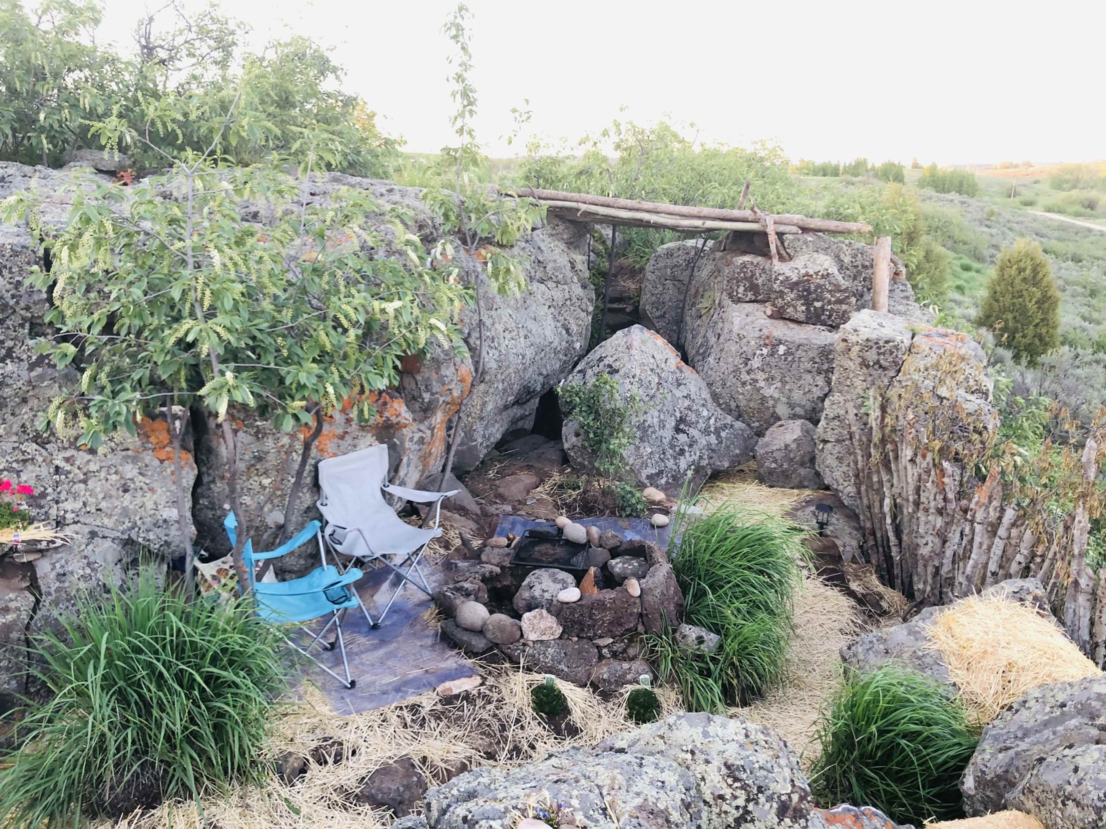 The scene features a small outdoor seating area with two folding chairs near a circular fire pit surrounded by large rocks and greenery.