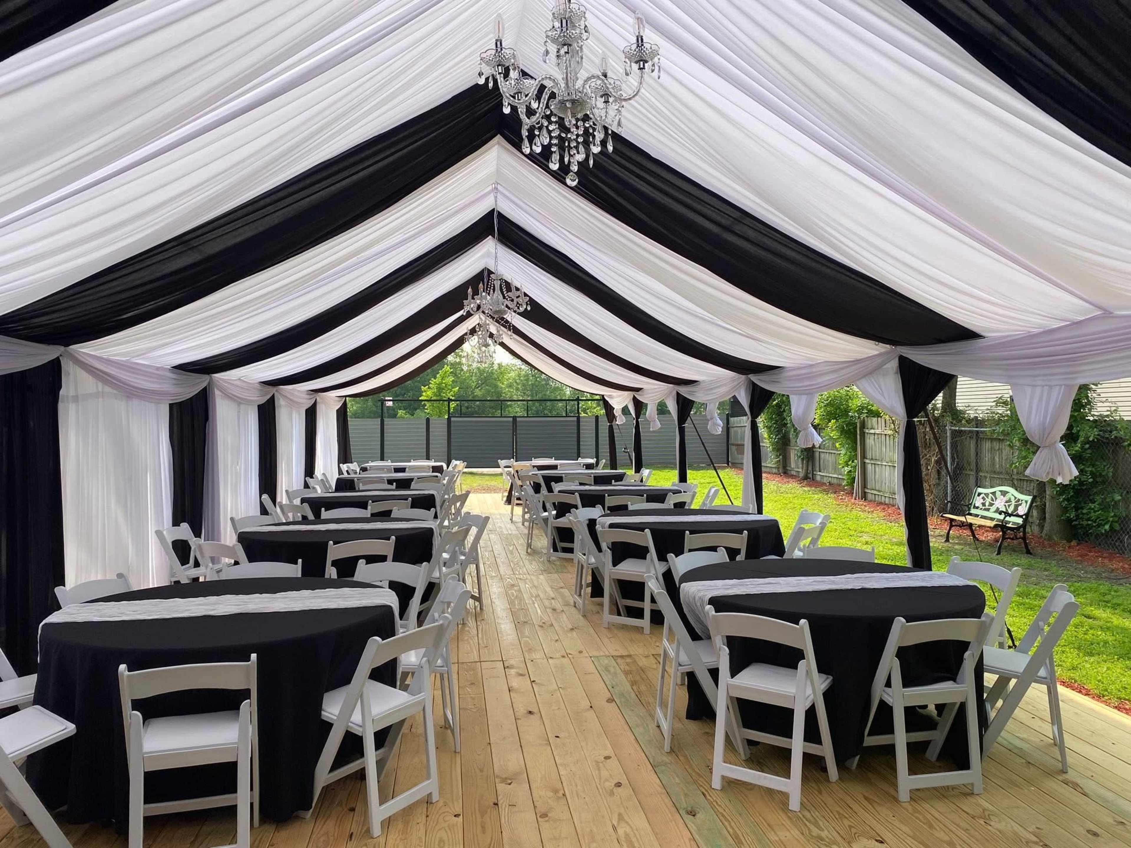 A large outdoor tent is set up with black and white draped fabric, featuring several tables with black linens arranged on a wooden floor.