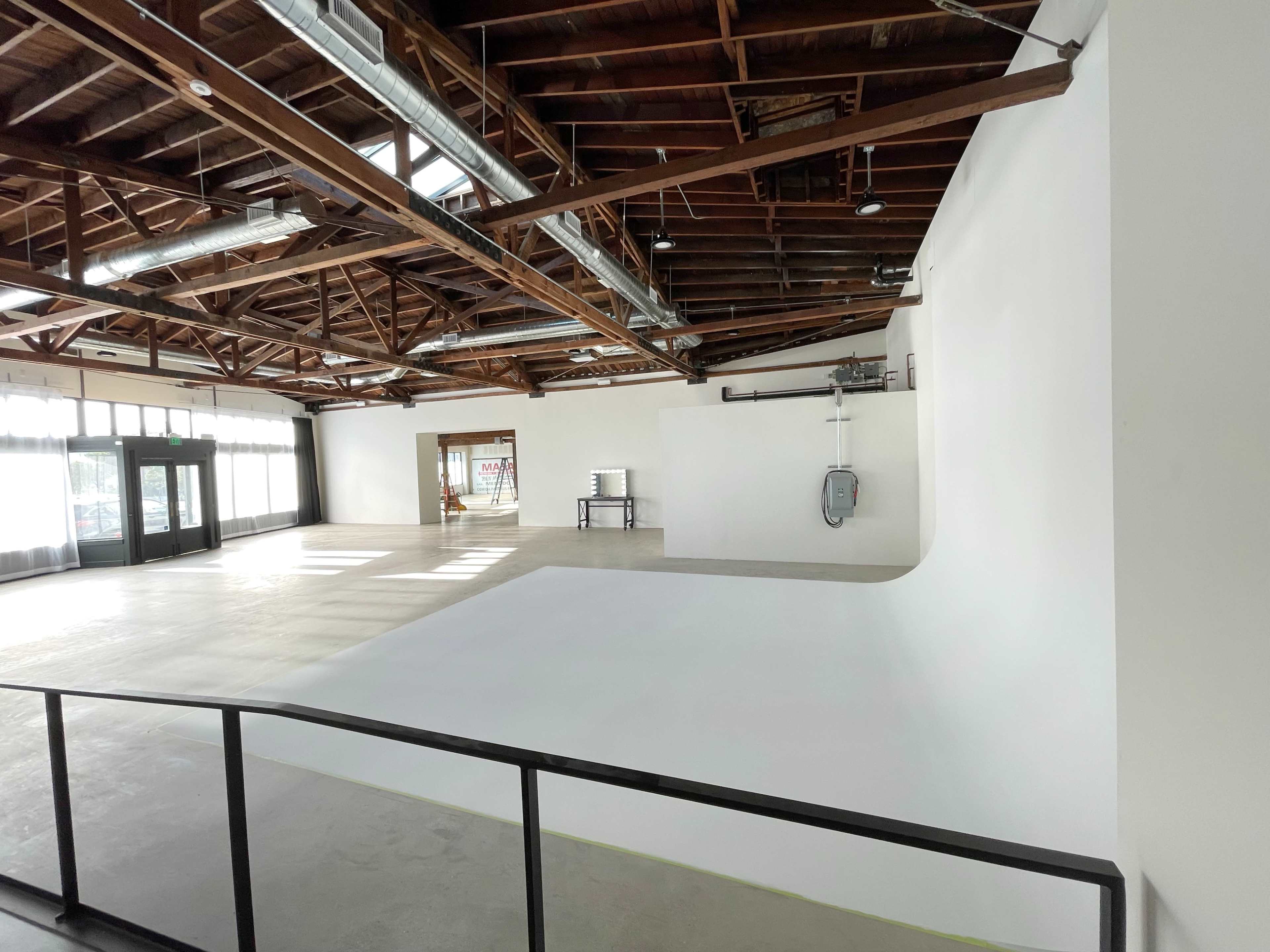 An empty interior space with a high, unfinished ceiling and large windows, featuring a seamless white wall at one end.