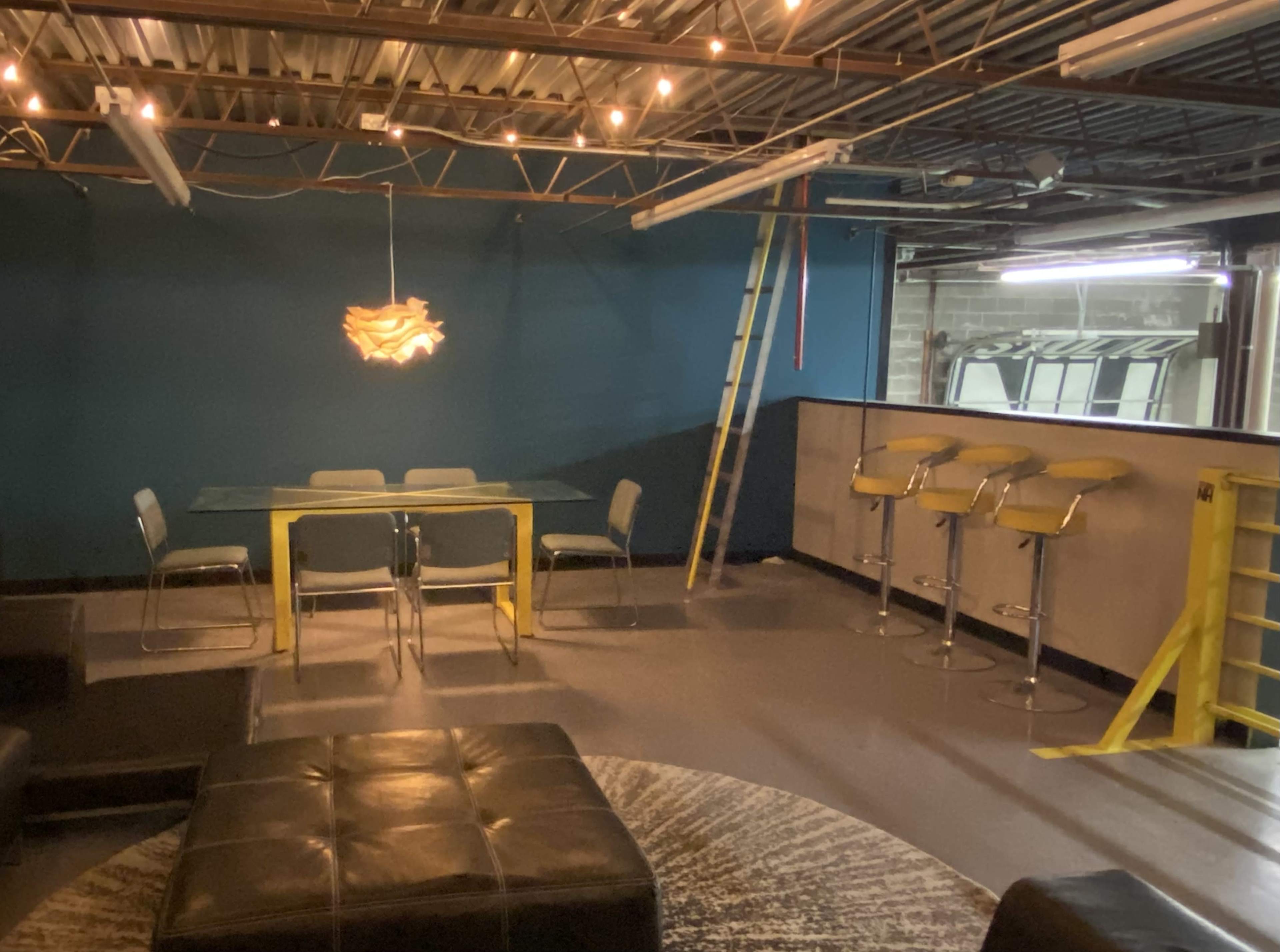 A sparsely furnished room with a glass dining table, several chairs, a bar with stools, and a ladder against a blue wall.