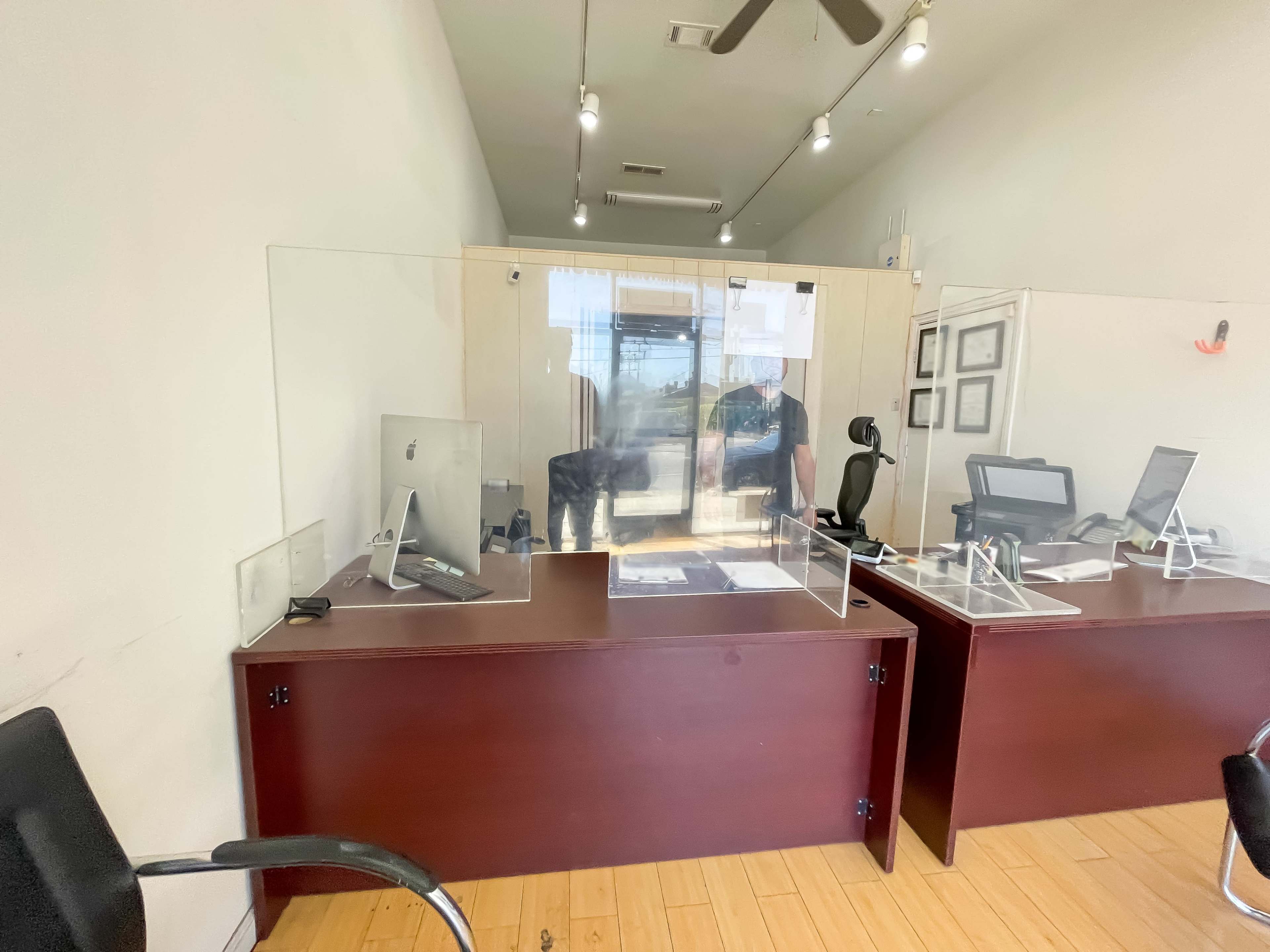 The image shows a small office space with two desks separated by clear acrylic partitions, and a large window at the back.