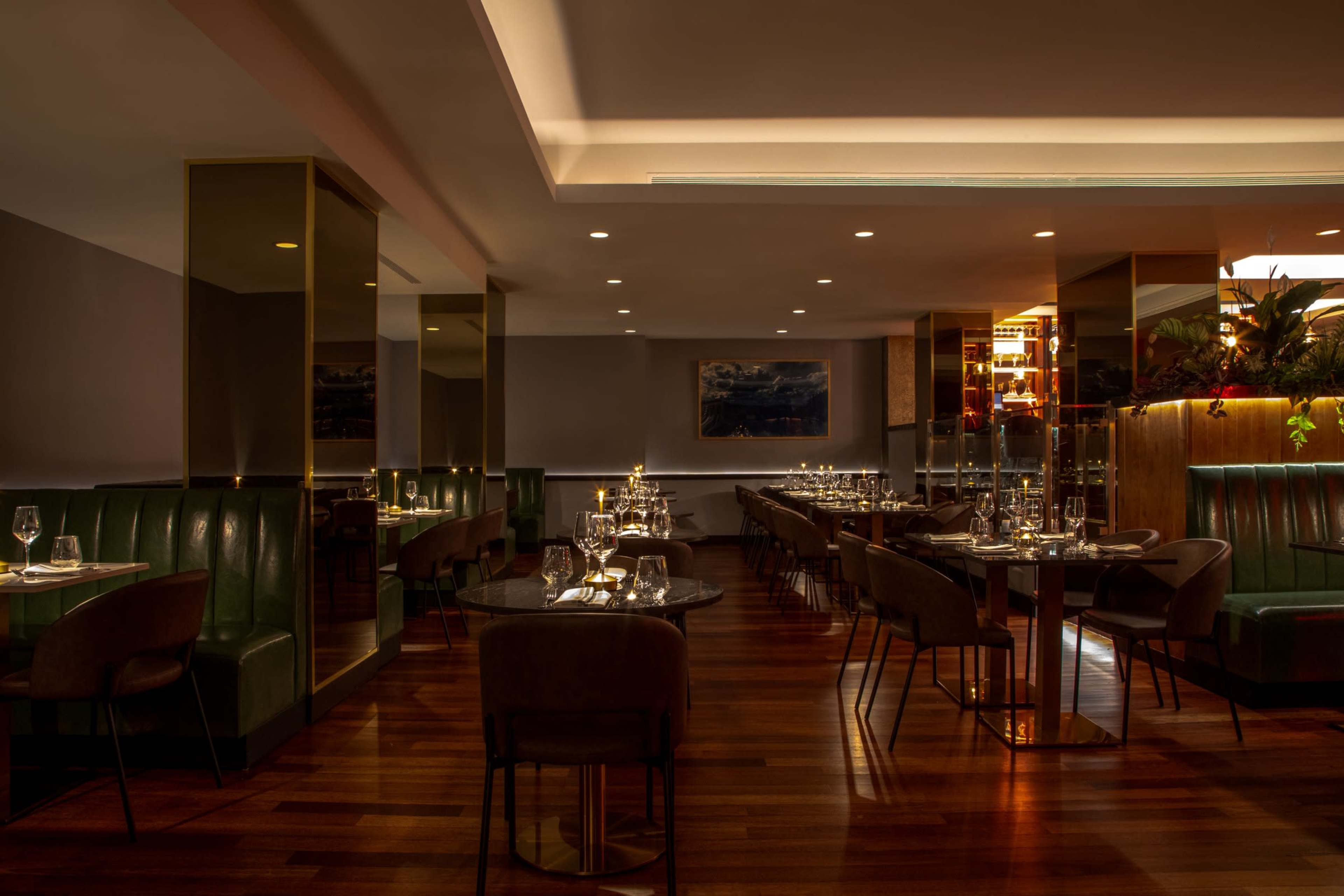 The image shows a modern restaurant with green booths, wooden floors, and softly lit tables set for dining.