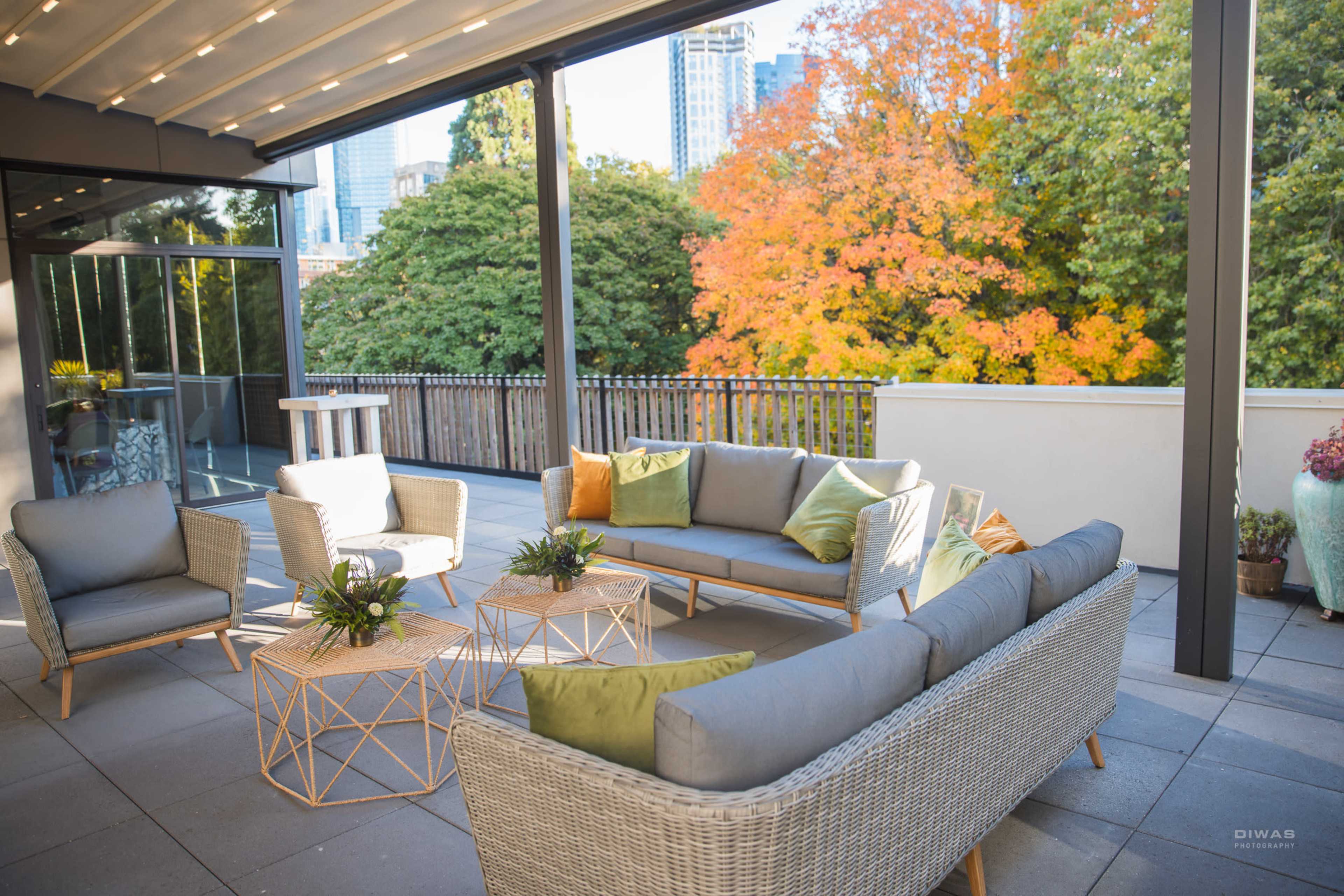 A modern outdoor seating area features wicker furniture with cushions and small tables, surrounded by greenery and autumn foliage in the background.