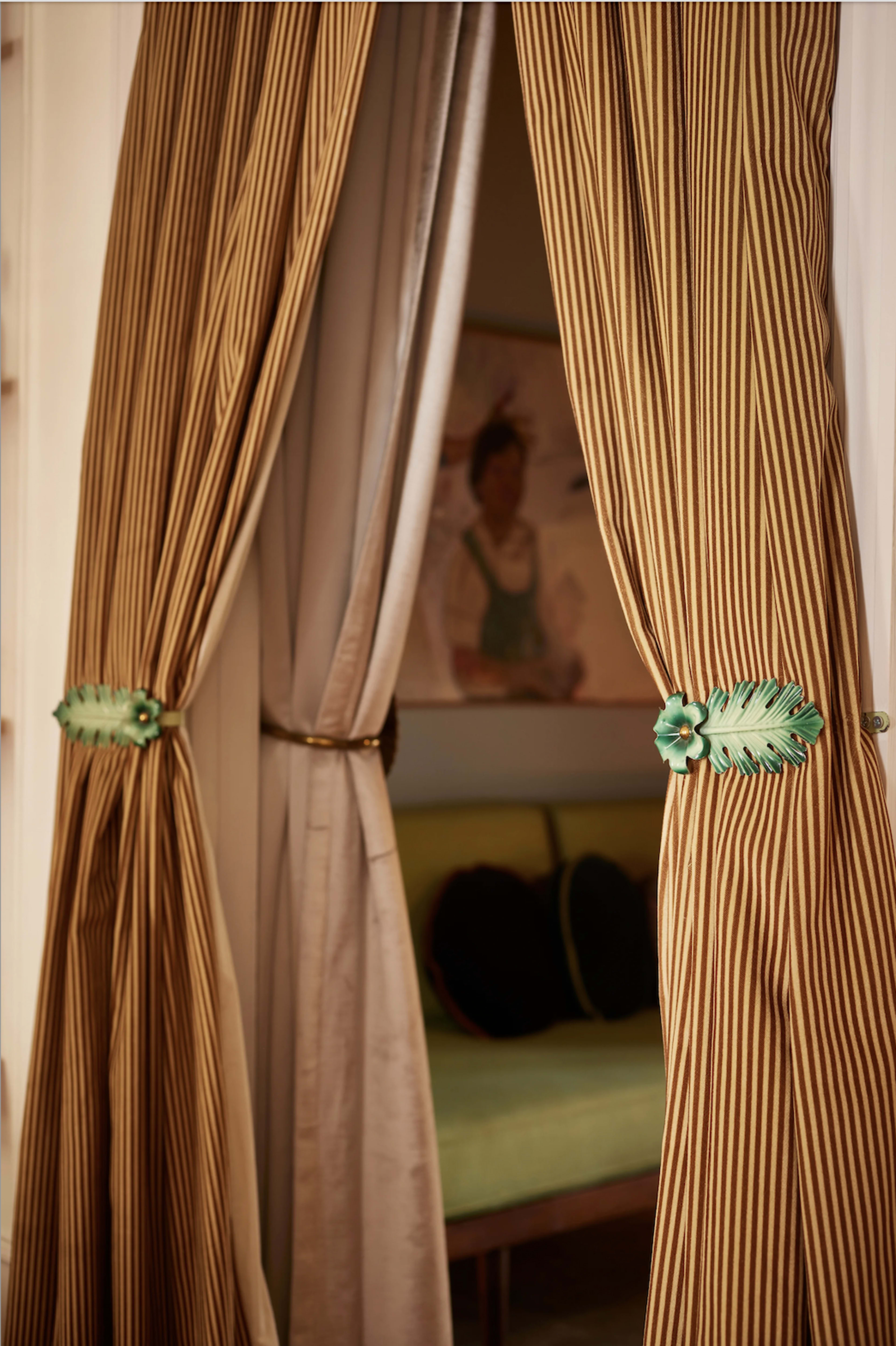 The image shows a pair of striped curtains with decorative floral tiebacks, partially revealing a brightly colored room in the background.