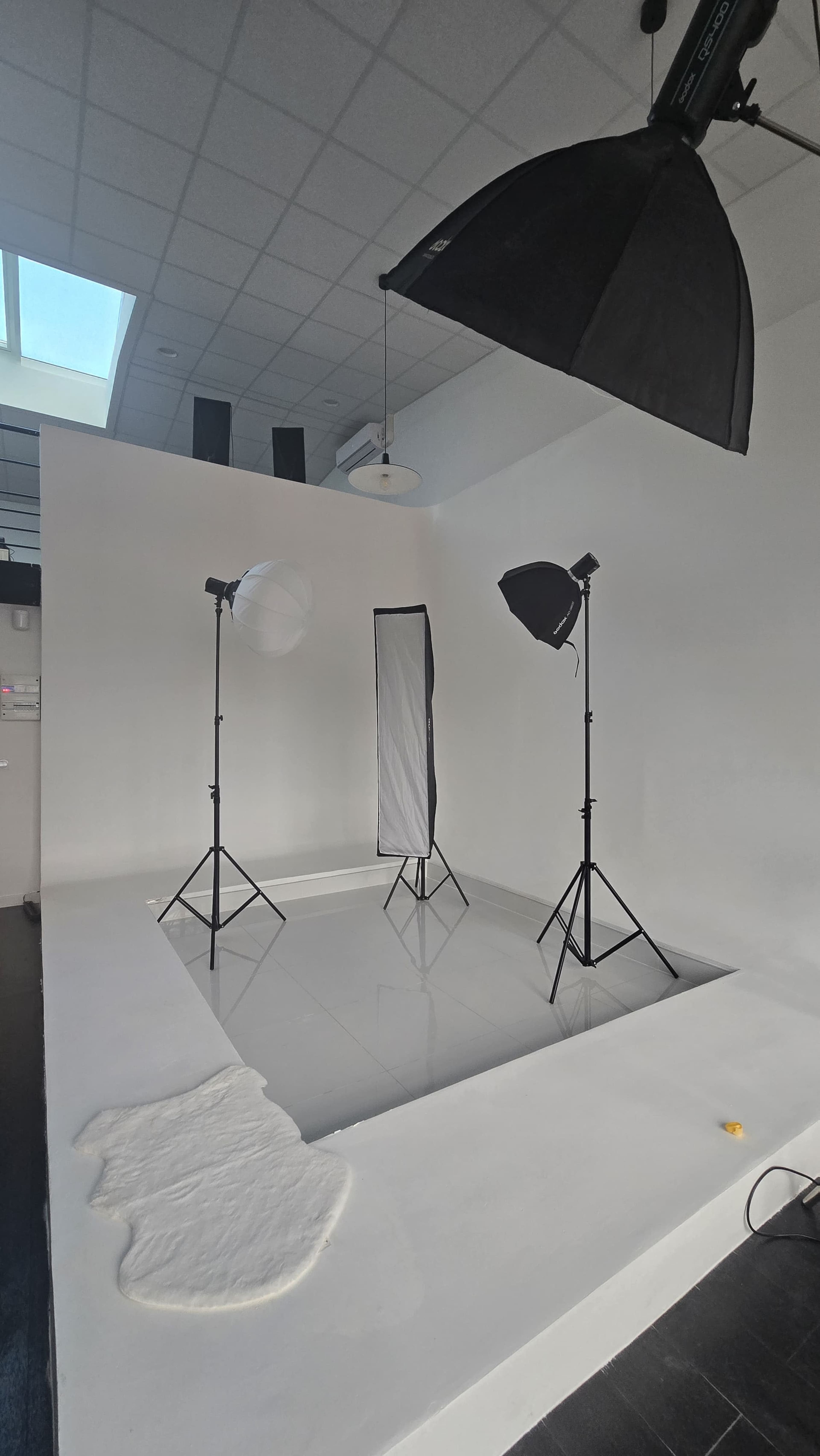 The image shows a photography studio with a large white backdrop, two softbox lights, and a reflective surface, set up for a photoshoot.