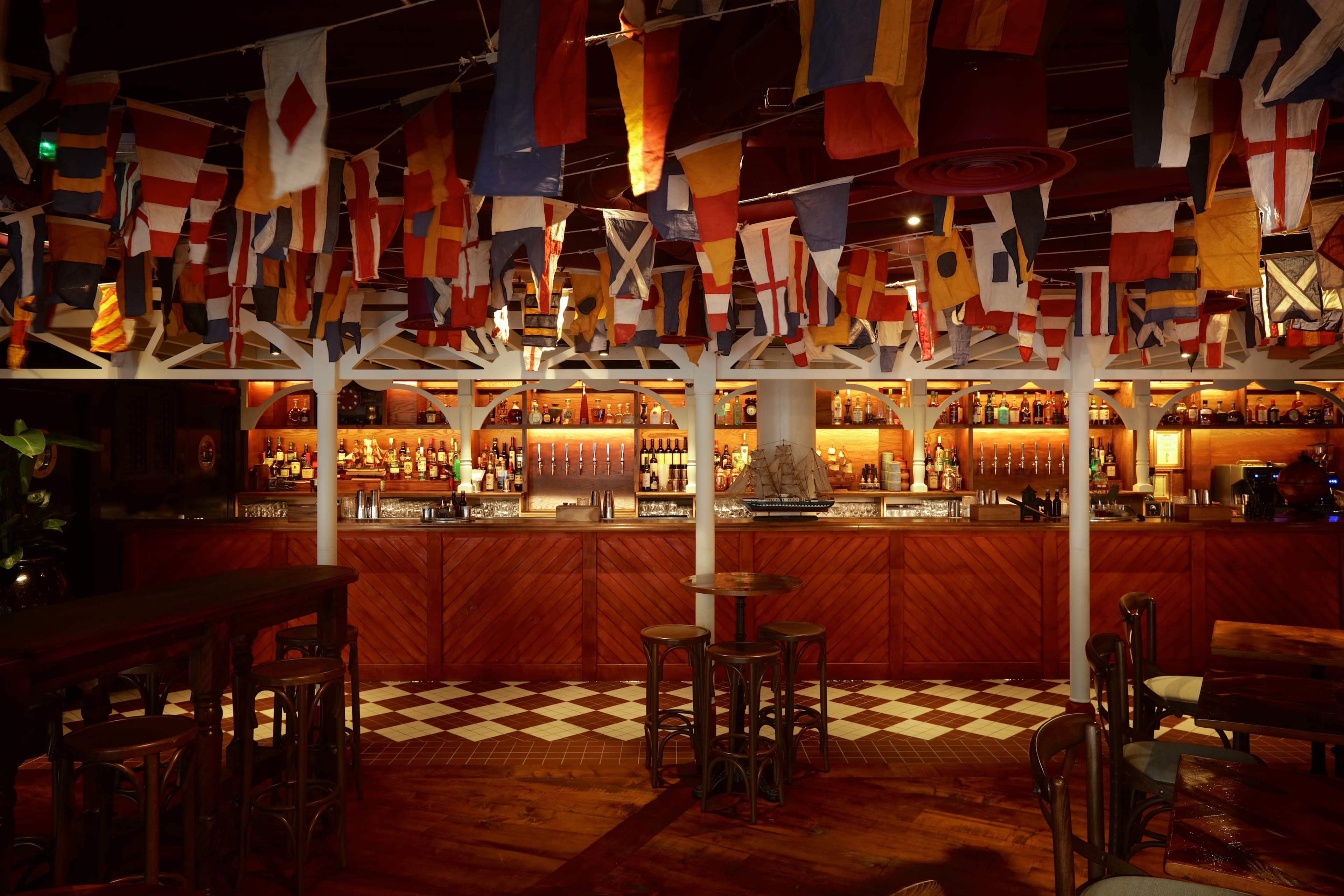 The image shows an interior of a bar adorned with colorful flags hanging from the ceiling and a well-stocked wooden bar in the background.
