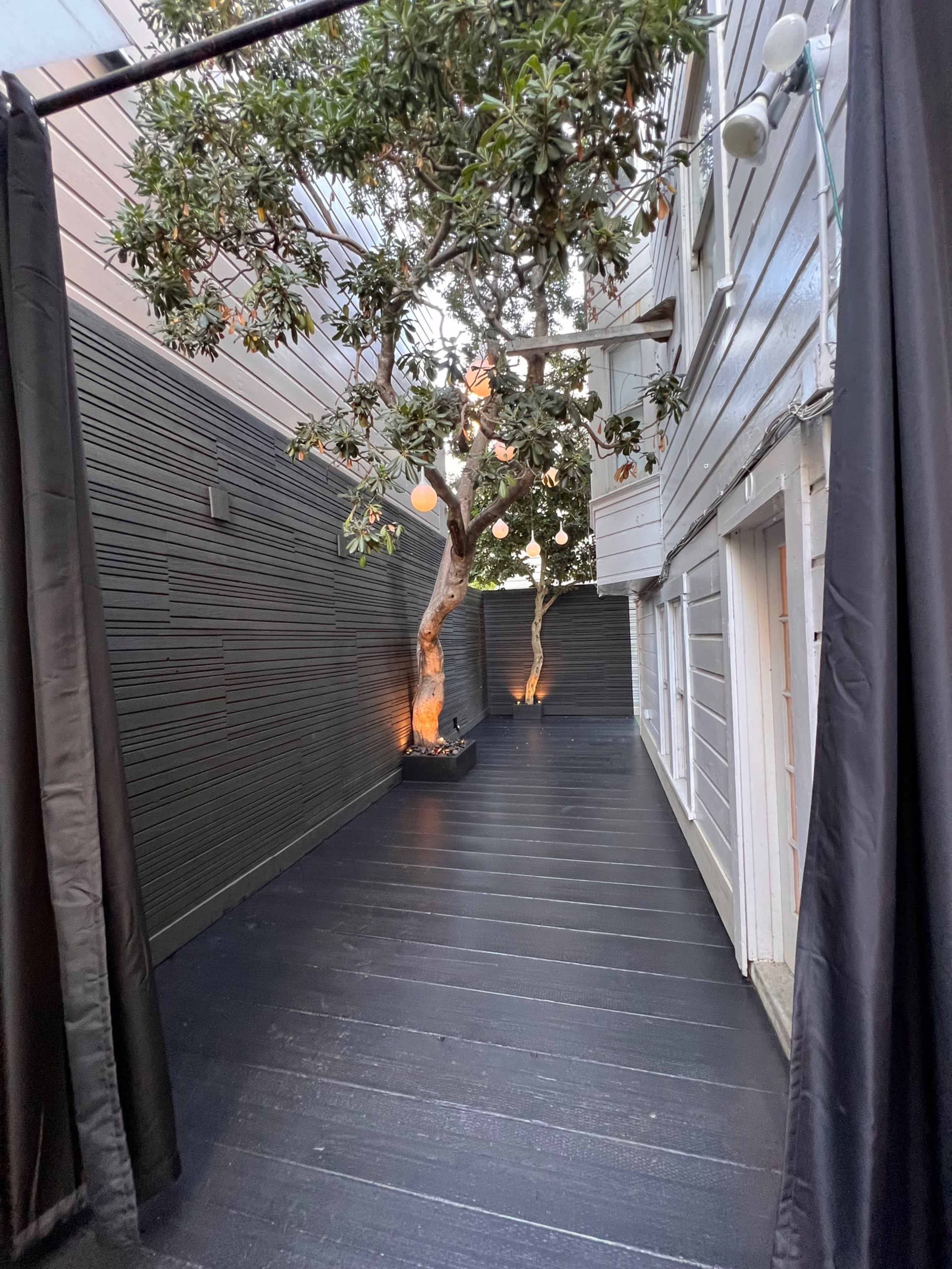 A narrow outdoor space with a wooden deck, flanked by walls, featuring a tree in the center and light fixtures above.