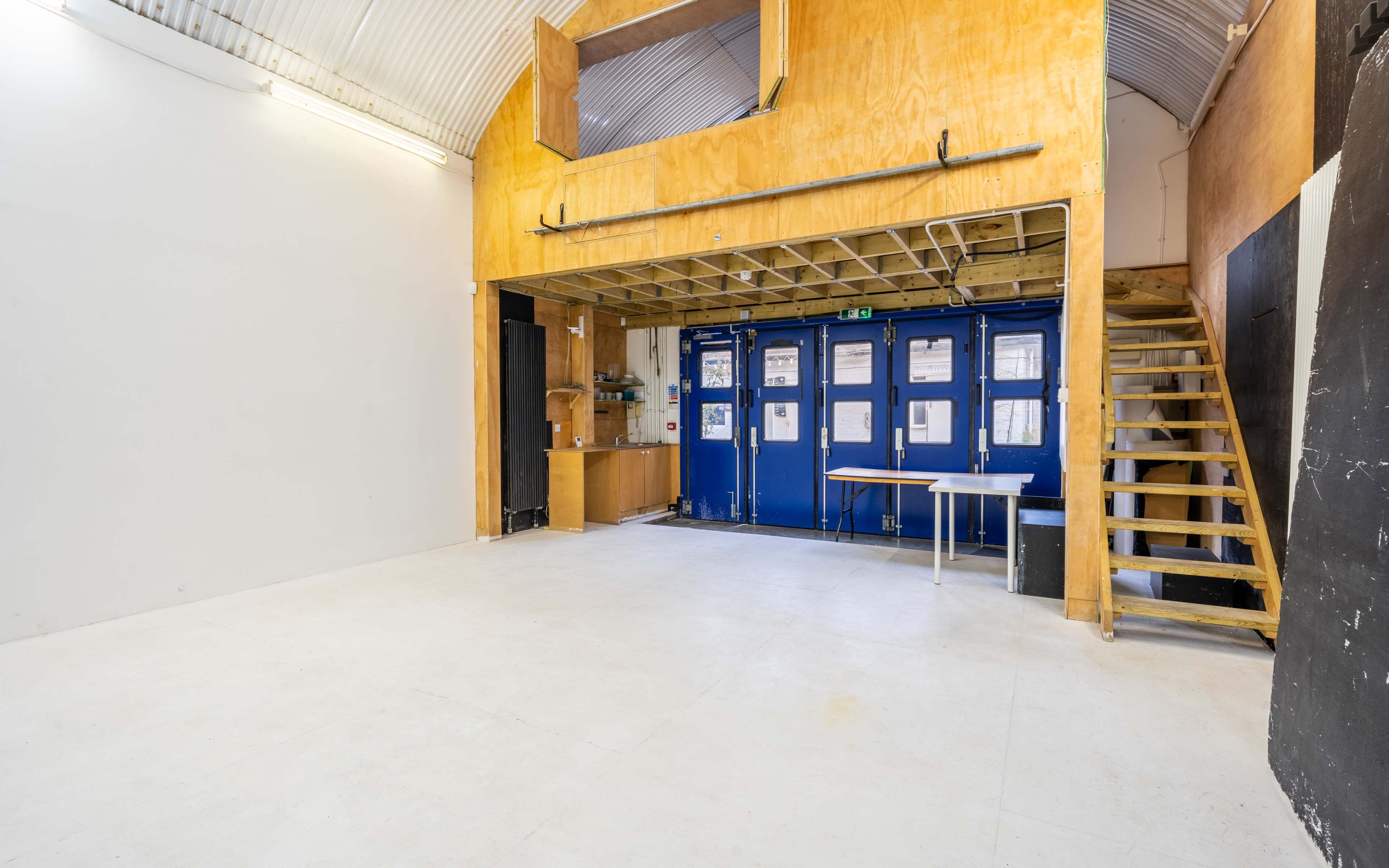 An empty interior space with a wooden loft area, blue doors, a staircase, and a table in the foreground.