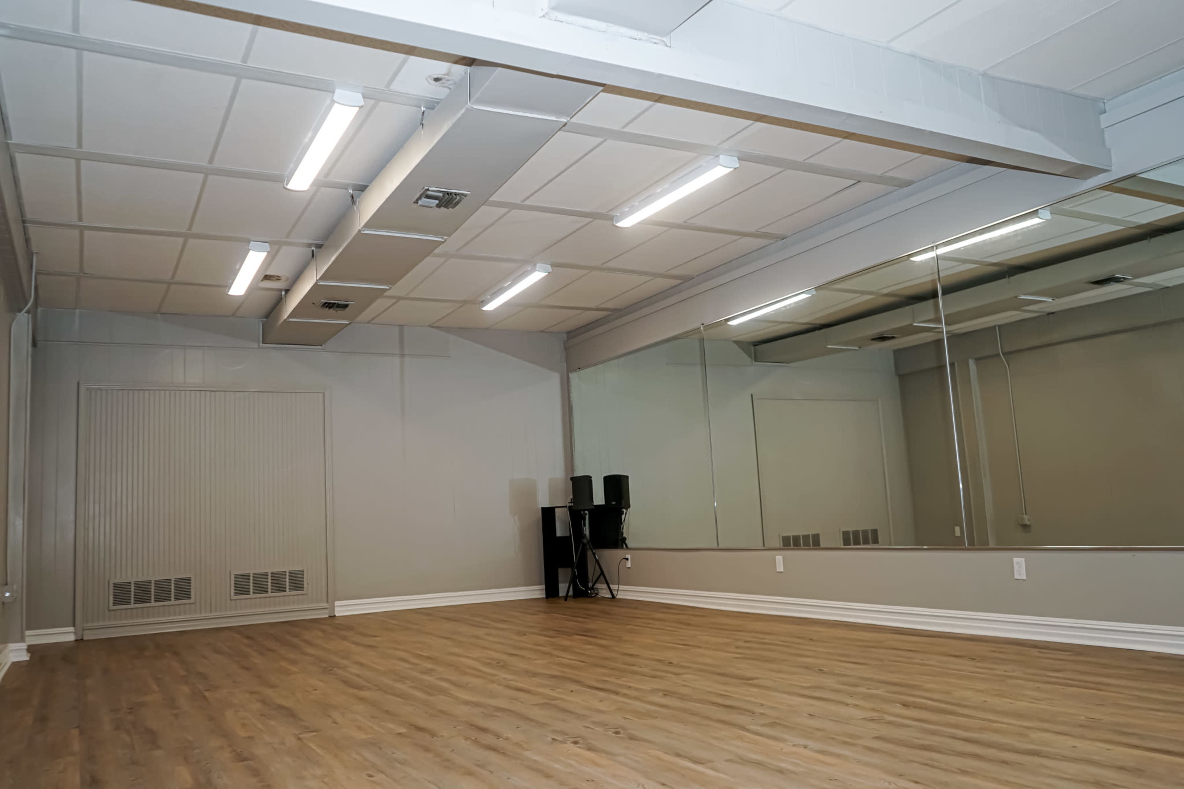 A spacious, empty room with wooden flooring, white walls, and large mirrors on one side, illuminated by overhead fluorescent lights.
